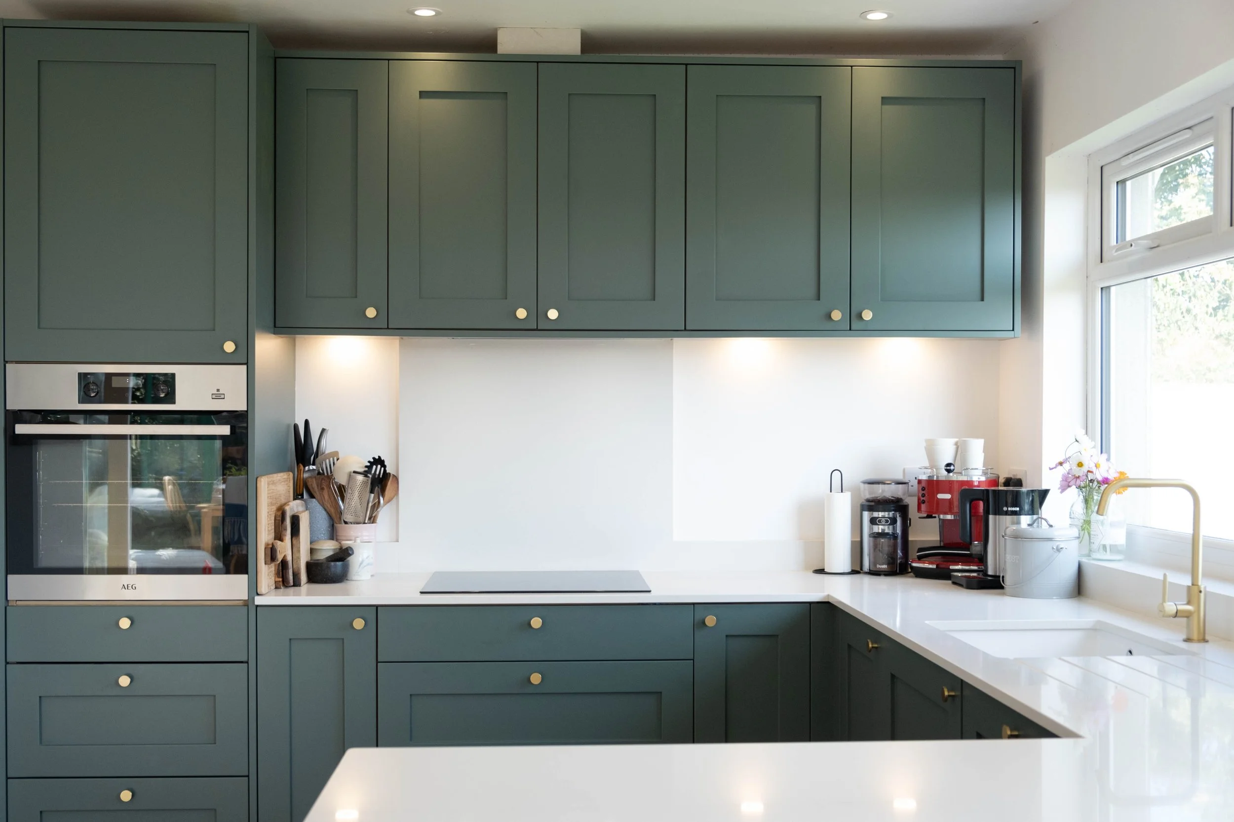 Modern kitchen with green cabinets, white countertops, stainless steel appliances, and various kitchen accessories near a window.