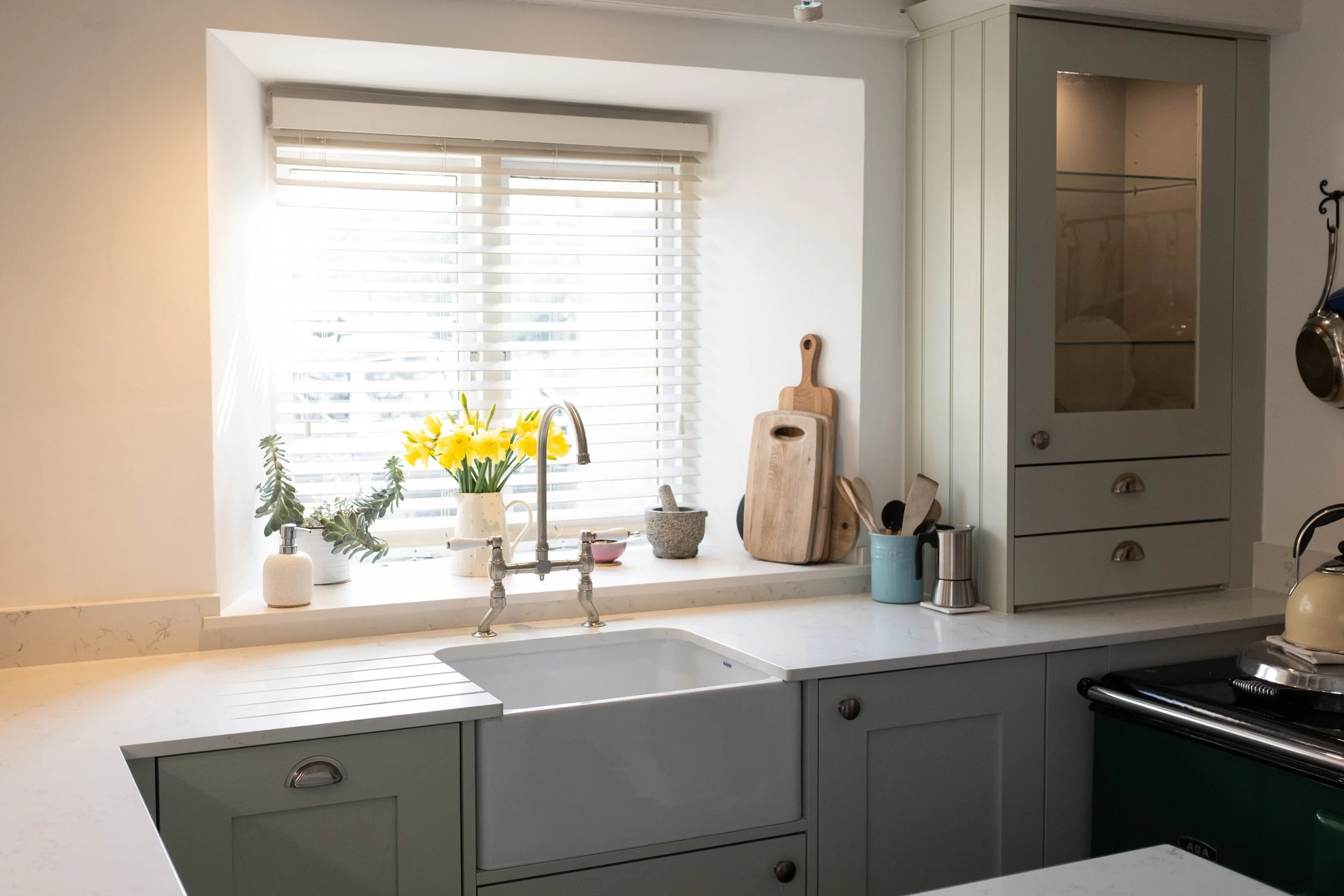 A kitchen with a white countertop, a window with blinds, and a gray cabinet. Decor includes a vase with yellow flowers, cutting boards, a mug, and kitchen utensils on the counter.