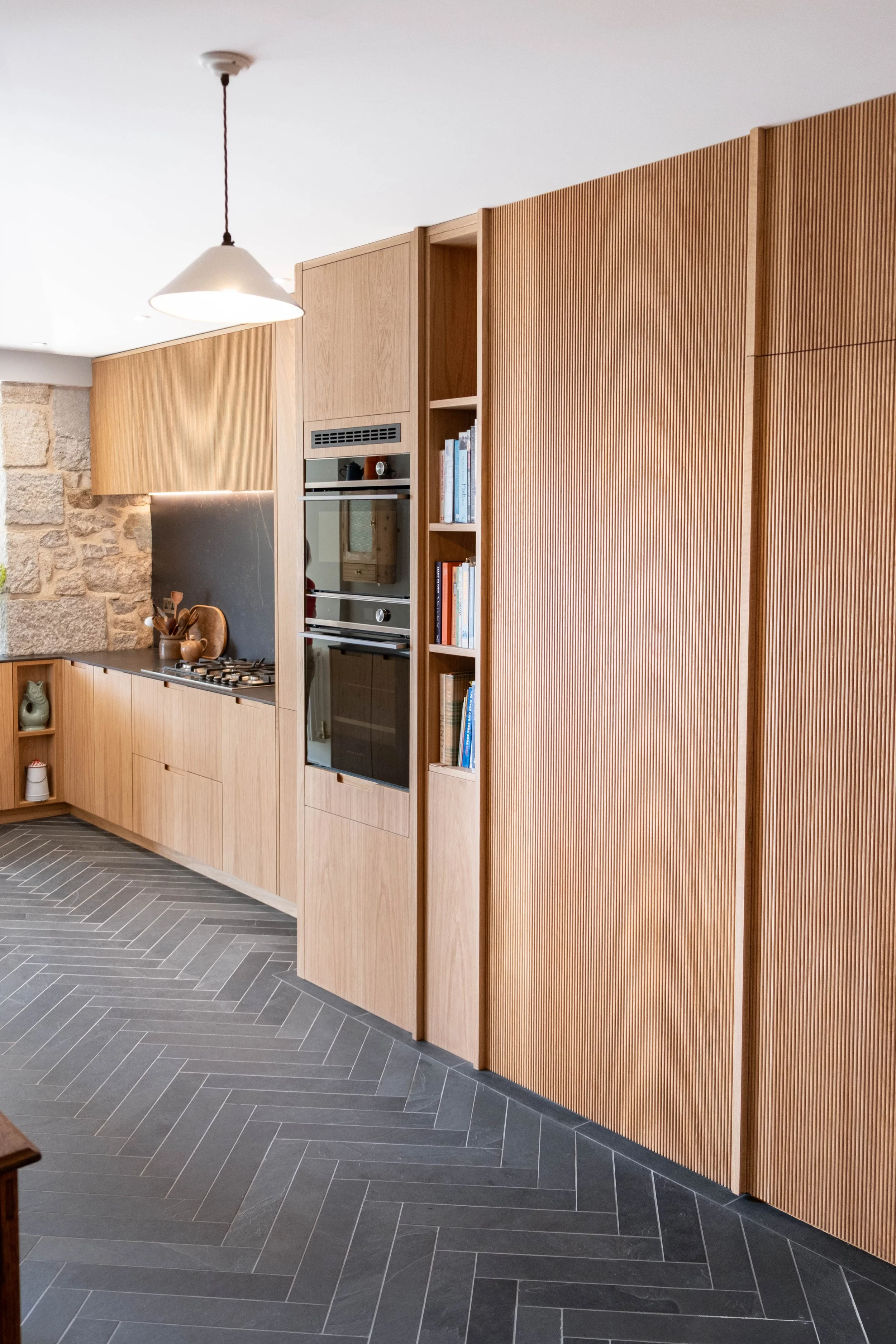 A modern kitchen with wooden cabinetry, a built-in oven, a gas stove, and a light fixture hanging from a white ceiling. The floor has dark gray herringbone tiles.