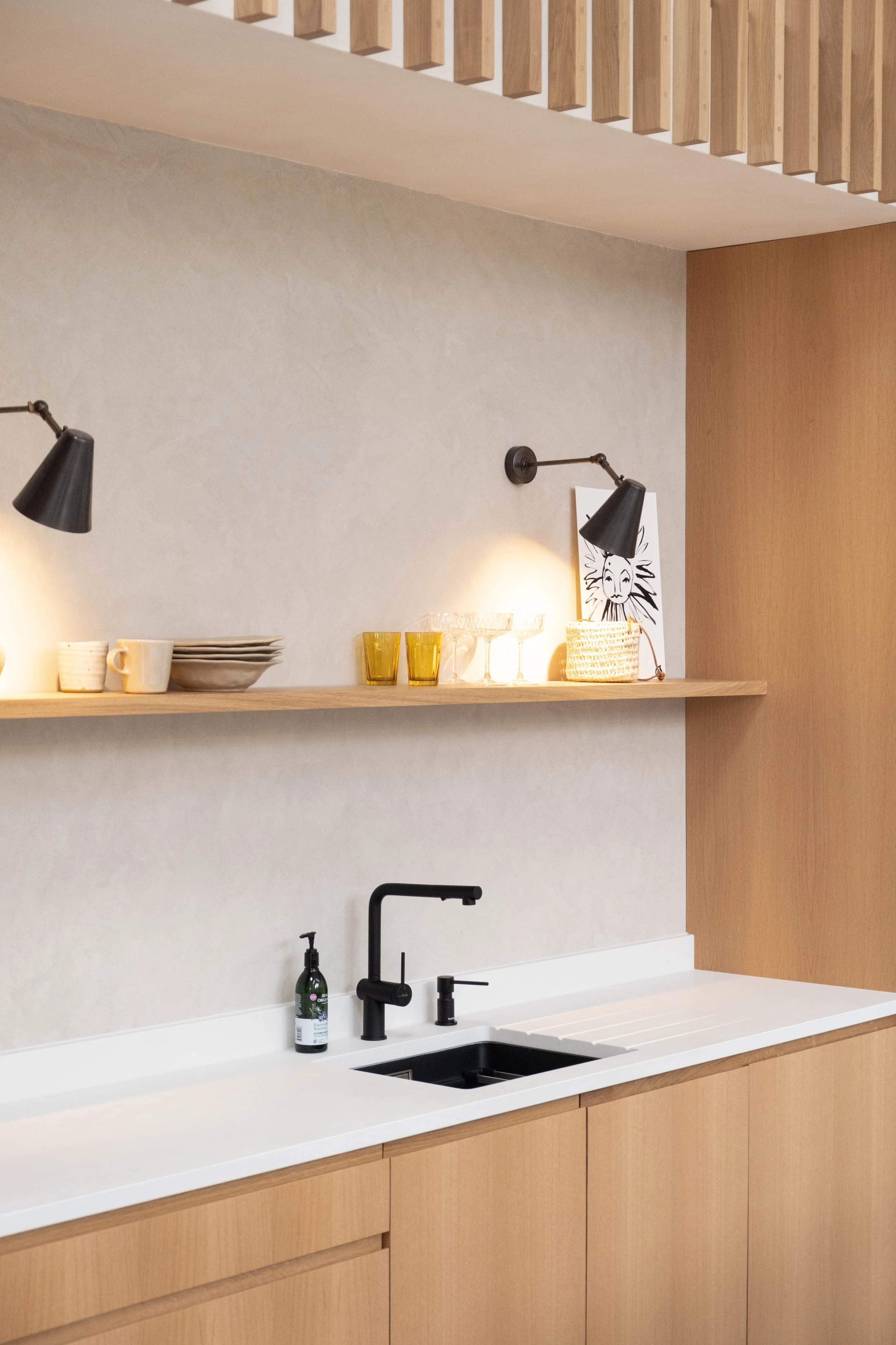 Kitchen with a white countertop, black faucet, soap dispenser, open shelves with white and yellow tableware, black wall-mounted lamps, a framed abstract face drawing, and wooden cabinetry.