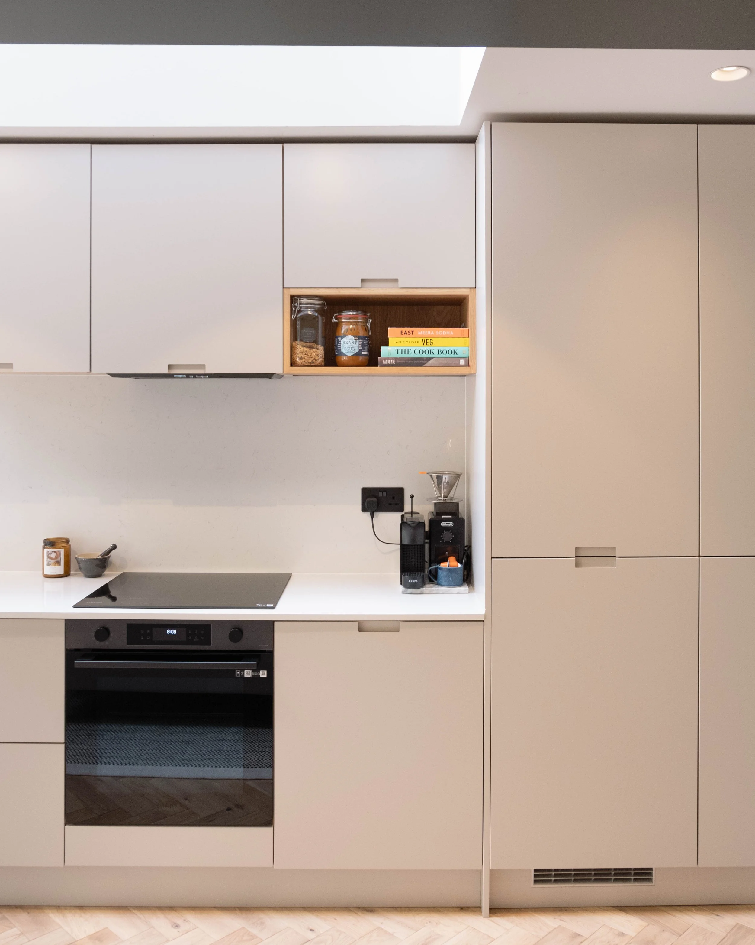 Modern kitchen with white cabinets, built-in oven, and a white countertop. There are some jars and books on a shelf above the counter. The kitchen has a clean and minimalist design.