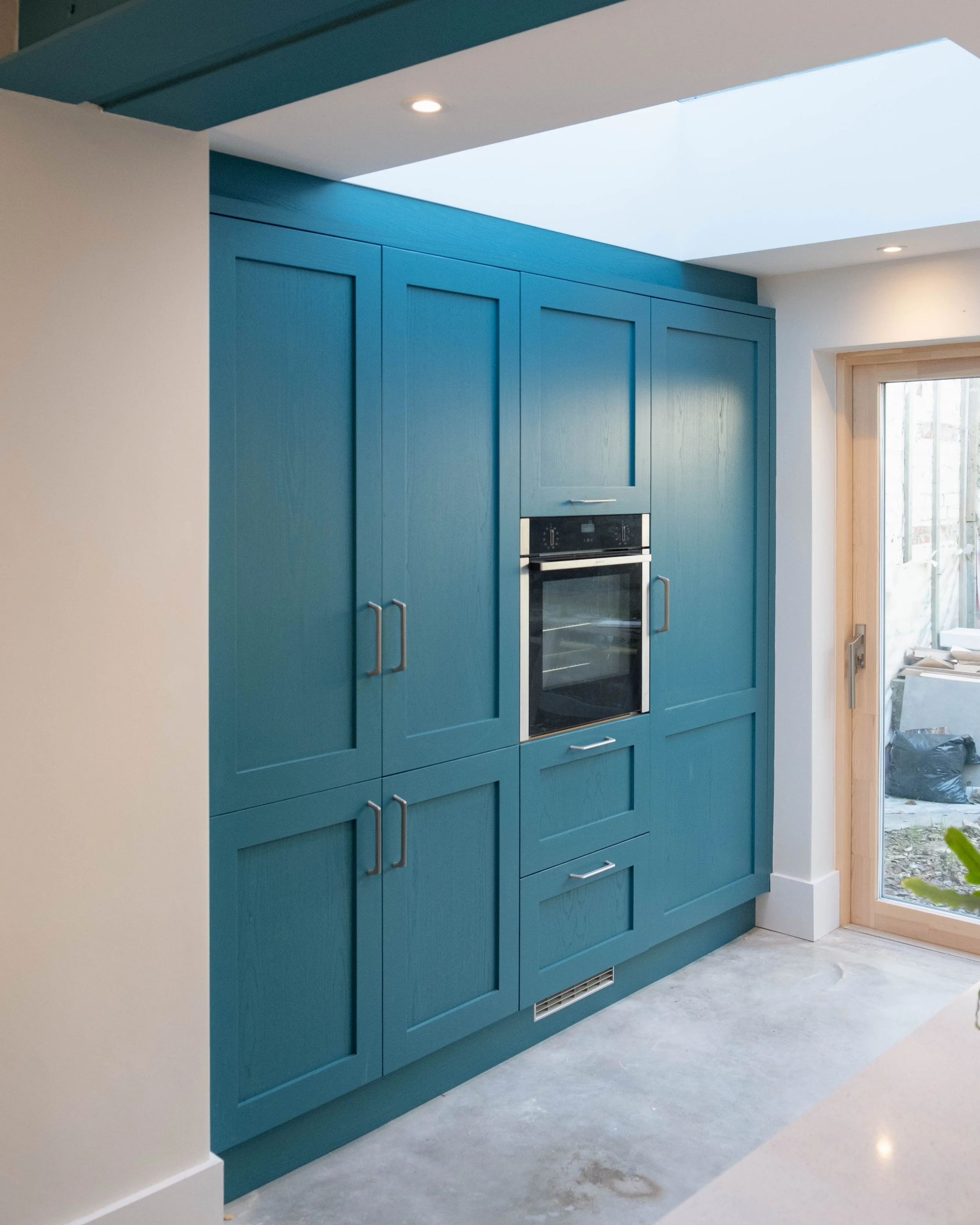 Interior view of a kitchen featuring blue cabinets with silver handles, a built-in double oven, and a large window letting in natural light.