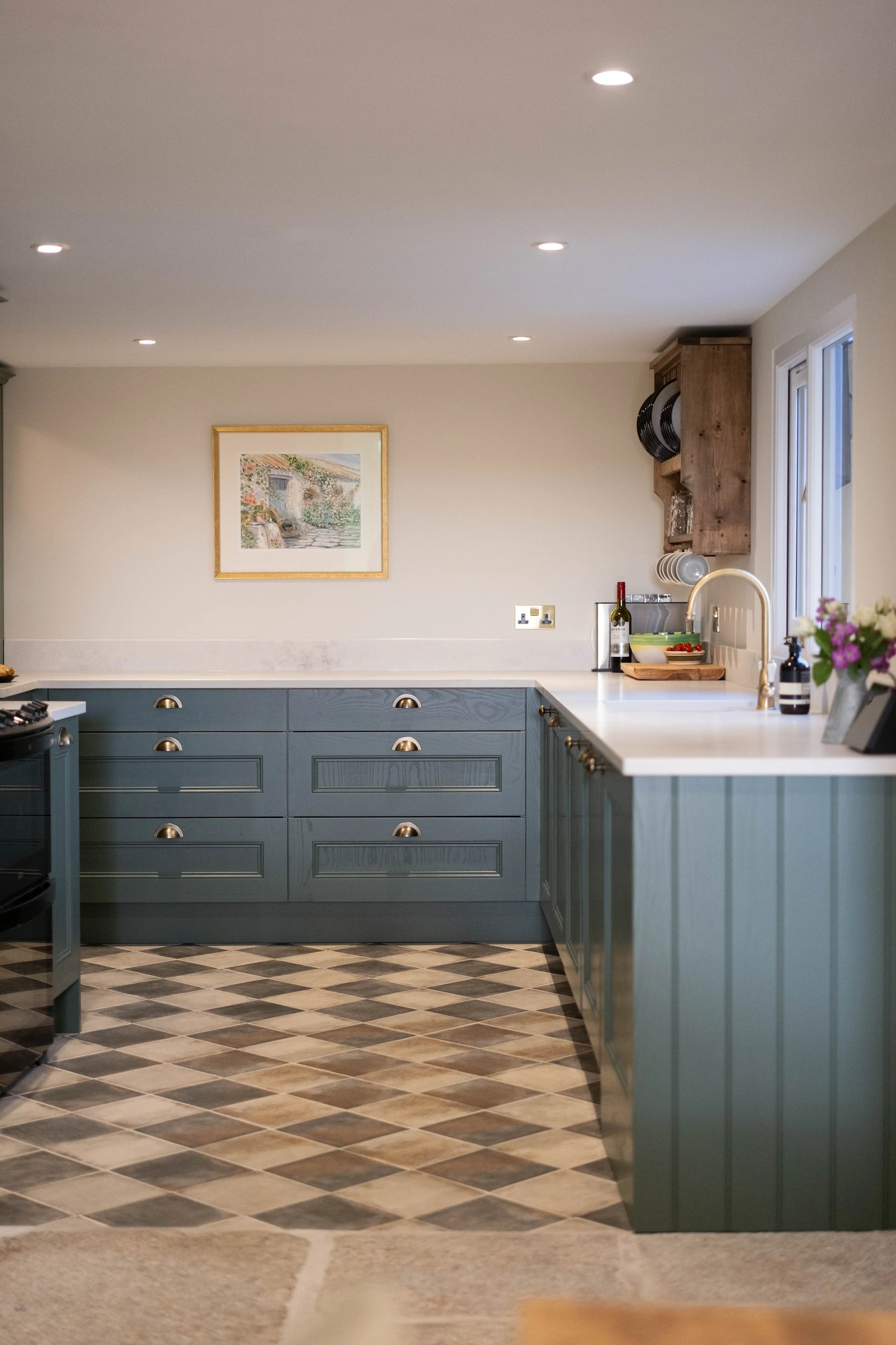 A kitchen with blue cabinets, a white countertop, hanging plates, a window, and a painting on the wall. The floor has a diamond patterned tile design.