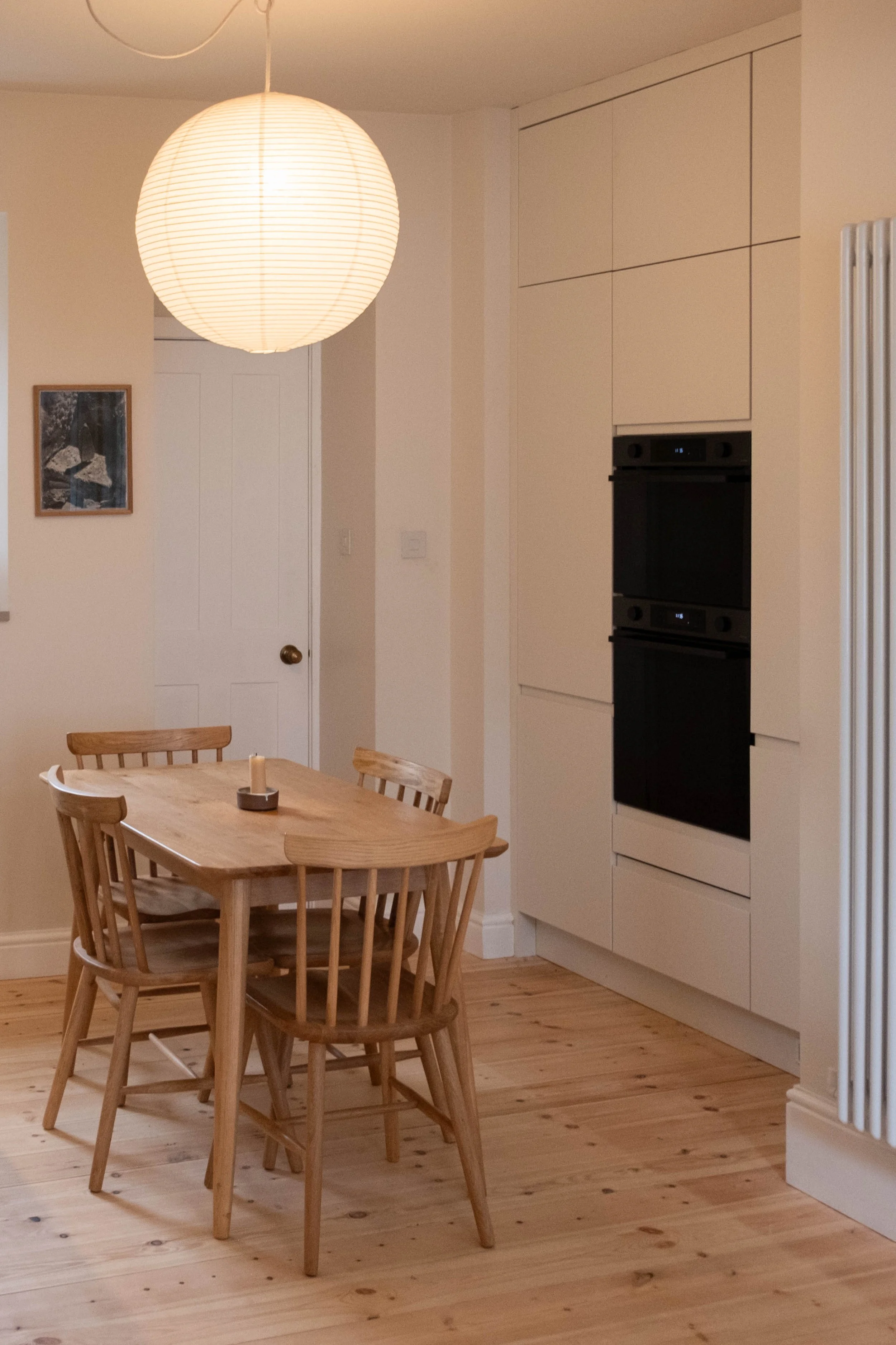 A wooden dining table with four matching chairs, a lit paper lantern hanging from the ceiling, and a kitchen with black built-in ovens and white cabinetry in the background.