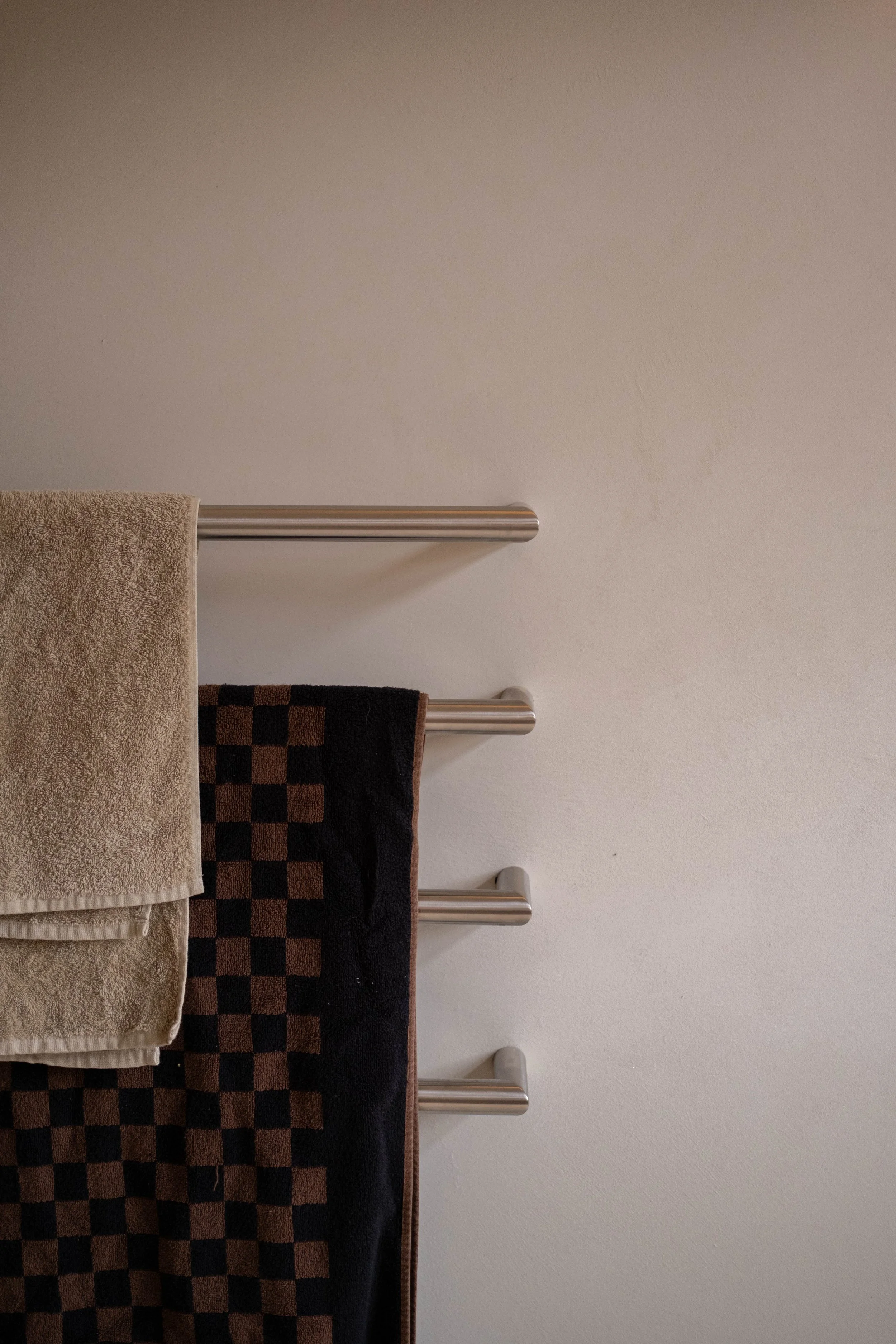 A wall-mounted towel rack with three towels hanging on it: a beige towel, a dark brown and orange checkered towel, and a plain black towel.