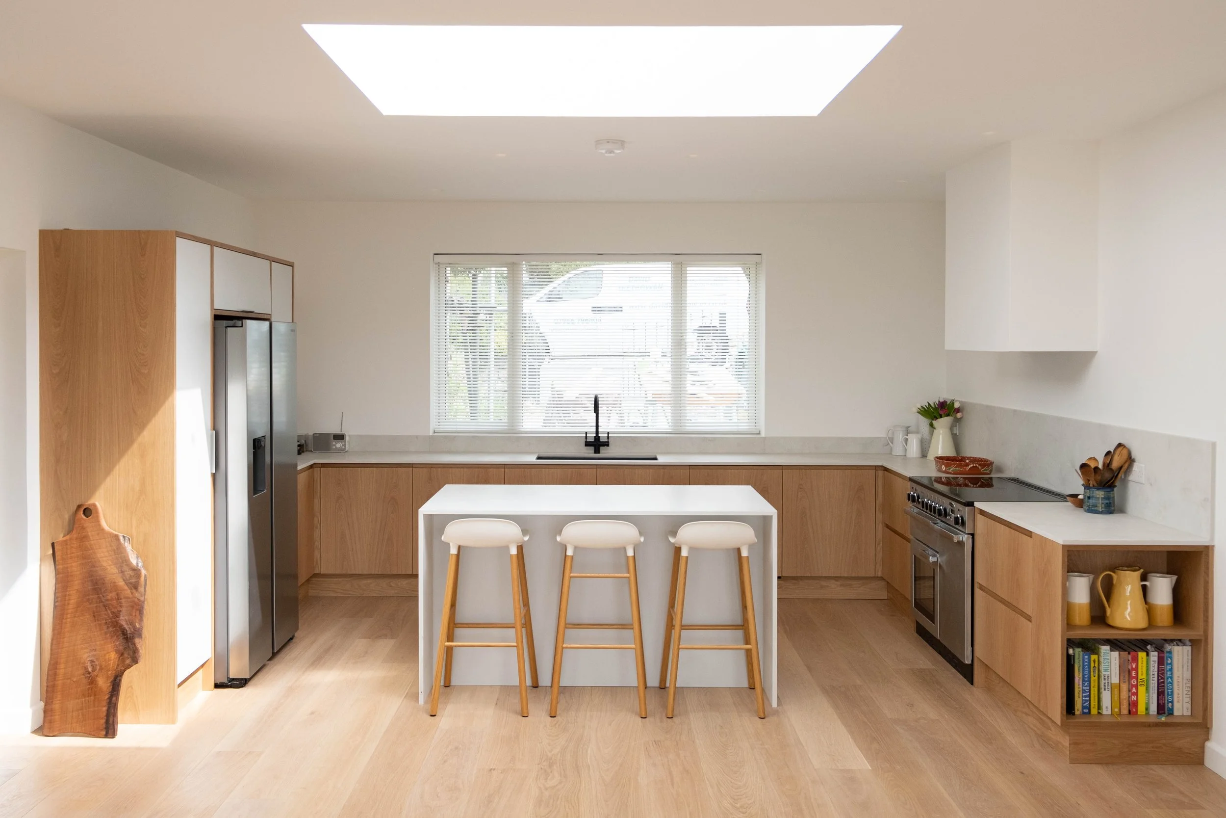 Modern kitchen with white cabinets, a white island with barstools, wooden flooring, a large window with blinds, stainless steel appliances, and decorative items on the counter.