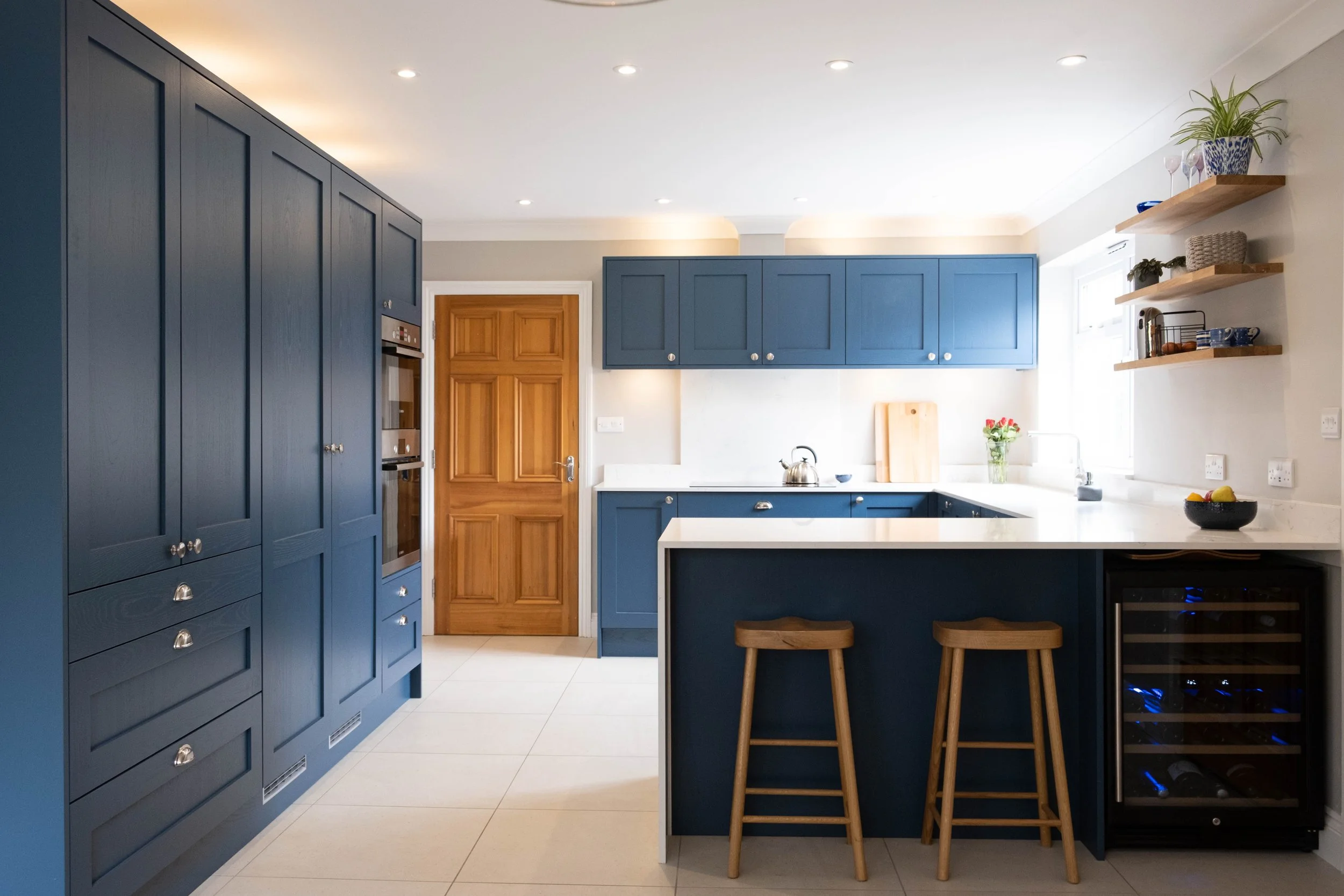Modern kitchen with navy blue cabinets, wooden door, open shelves with plant and dishes, island with two wooden stools, white countertops, and a window with sunlight.