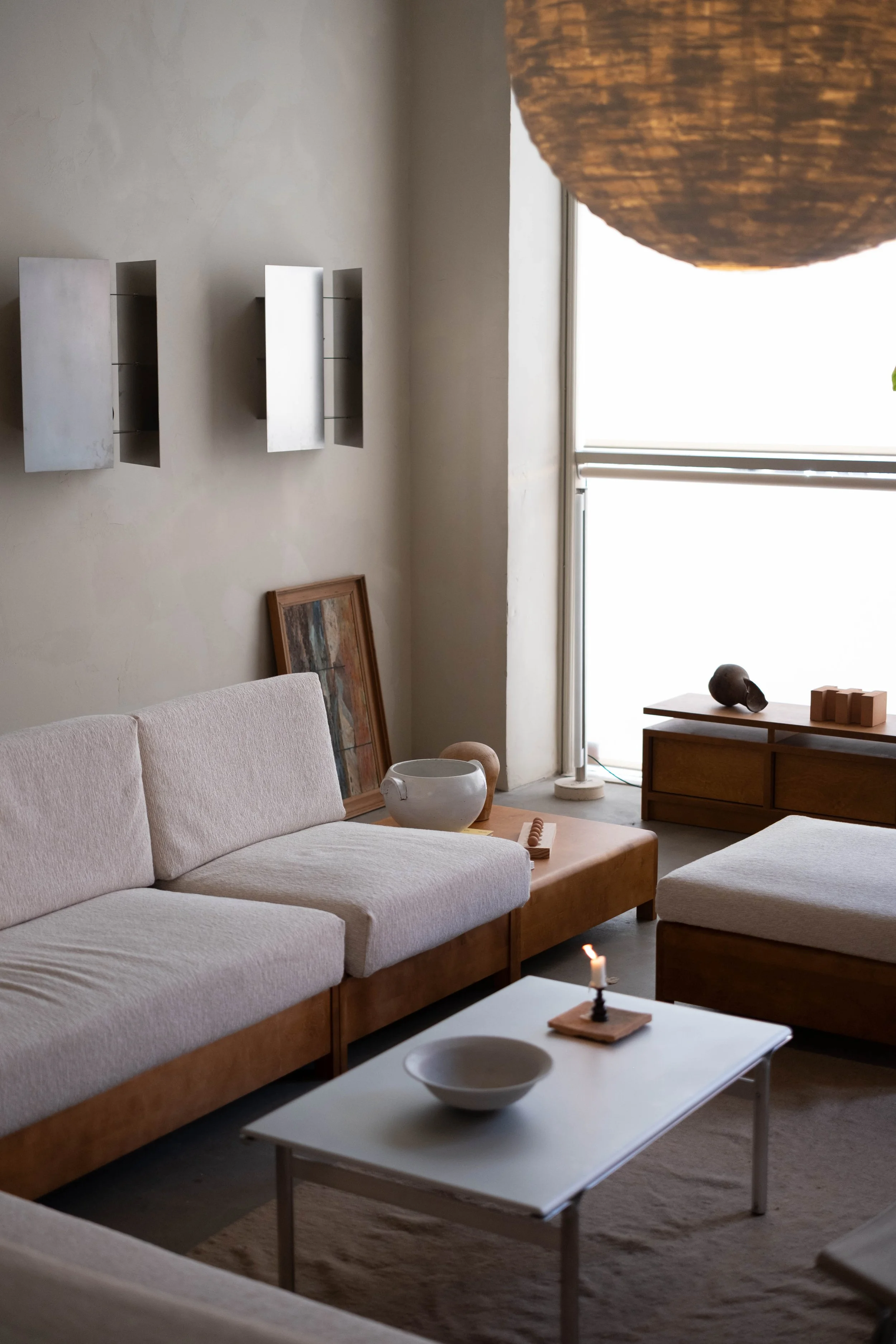 Living room with cream-colored sofa, wooden coffee table, decorative objects, painting, white walls, and large window letting in natural light.