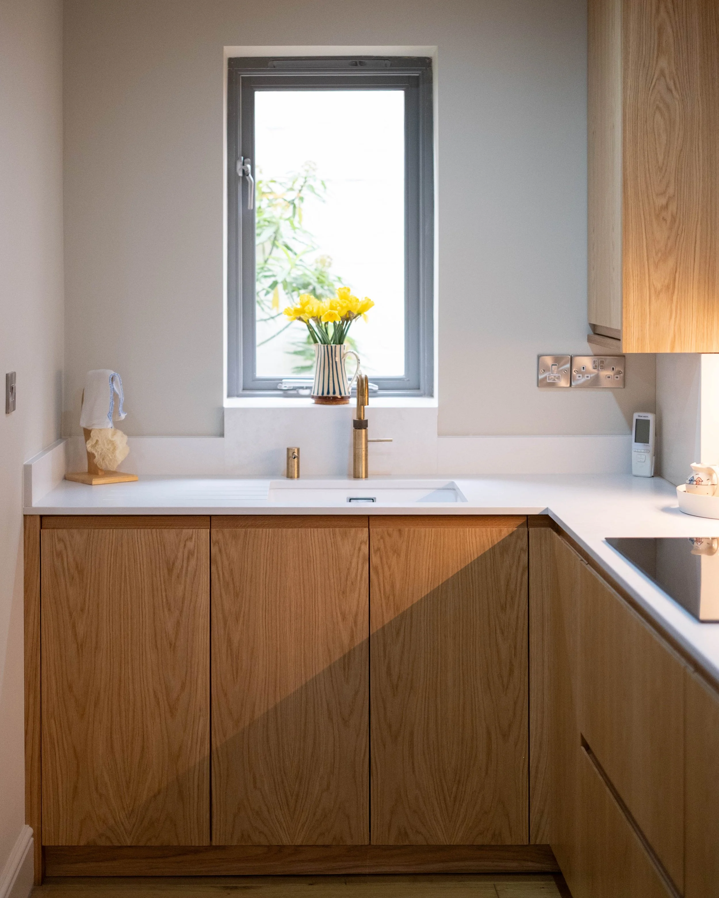 Kitchen with wooden cabinets, white countertop, a window with a vase of yellow flowers, and a gold faucet.