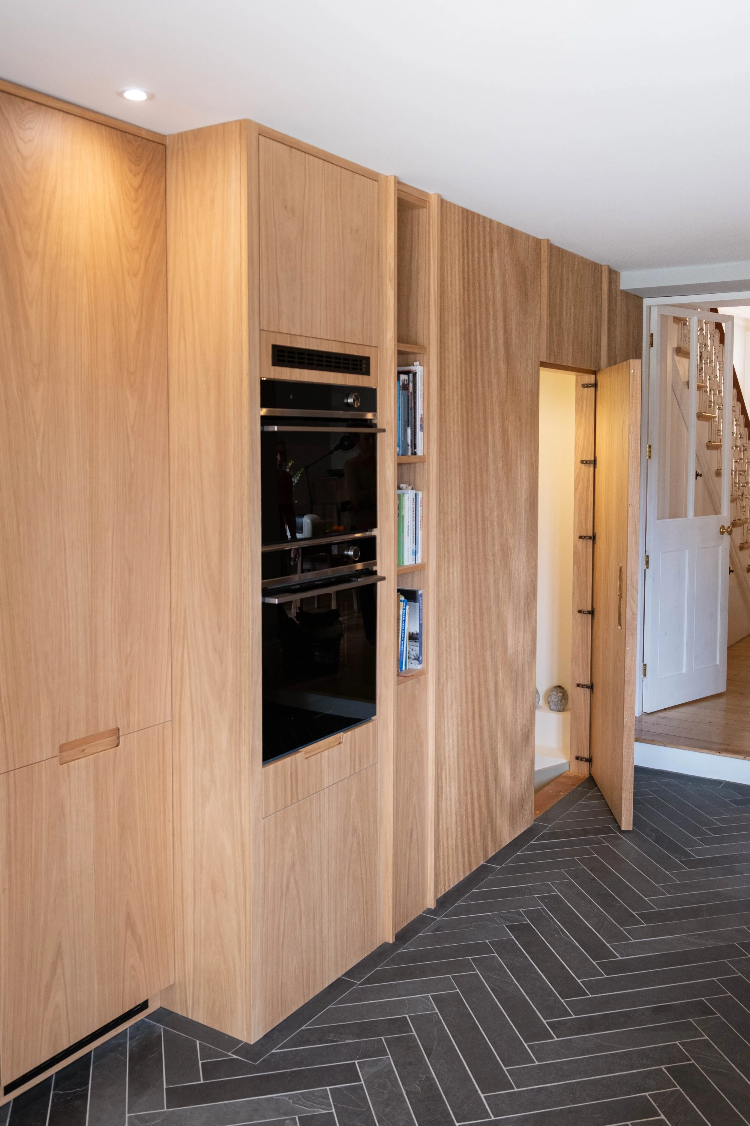 Interior view of a modern kitchen with light wood cabinets, a built-in oven, open shelving, and a doorway leading to a staircase. The floor has black and gray herringbone tiles.