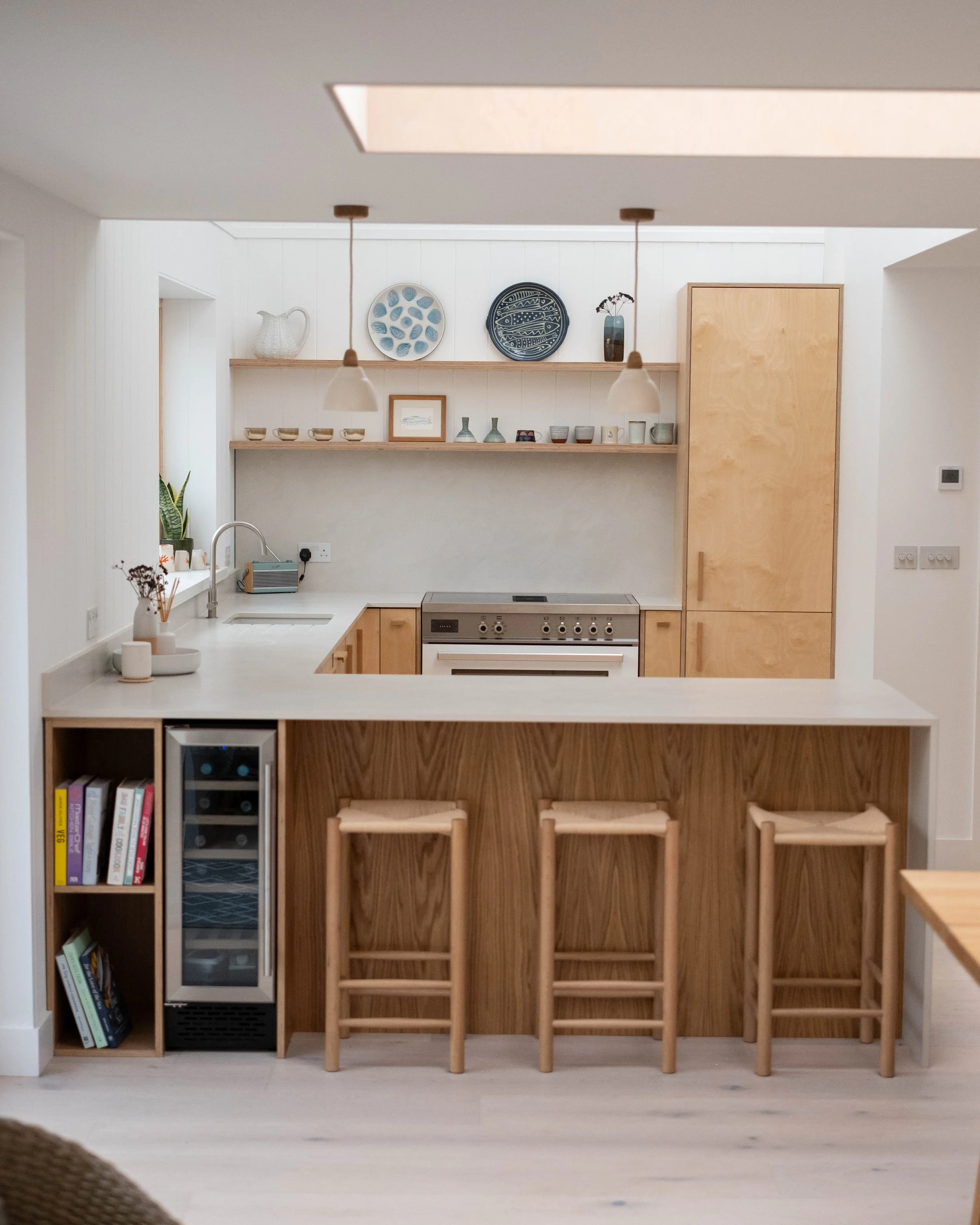 A kitchen with a white countertop, a wooden breakfast bar with three wooden stools, open shelving with dishes and decorative items, a stainless steel oven, a tall wooden cabinet, a small wine cooler, and some books on a side shelf.