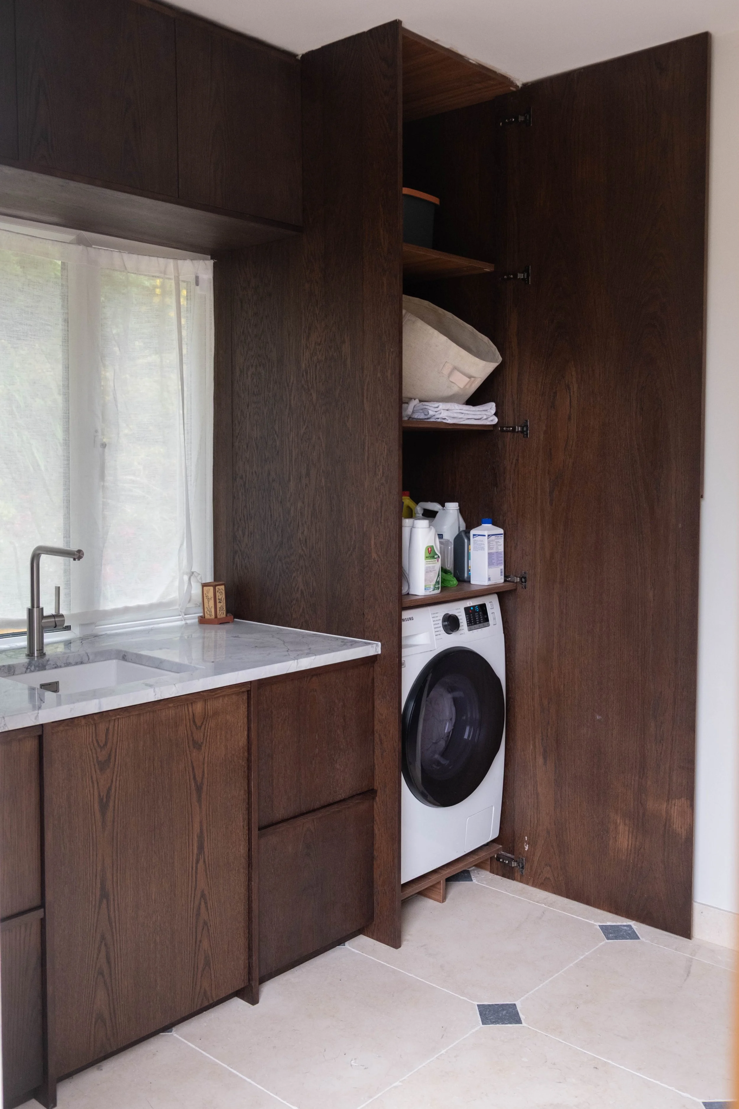 Kitchen with dark wood cabinets, a white marble countertop, a stainless steel sink, window with white curtains, and a front-loading washing machine. Storage of laundry items and a basket inside an open cabinet.