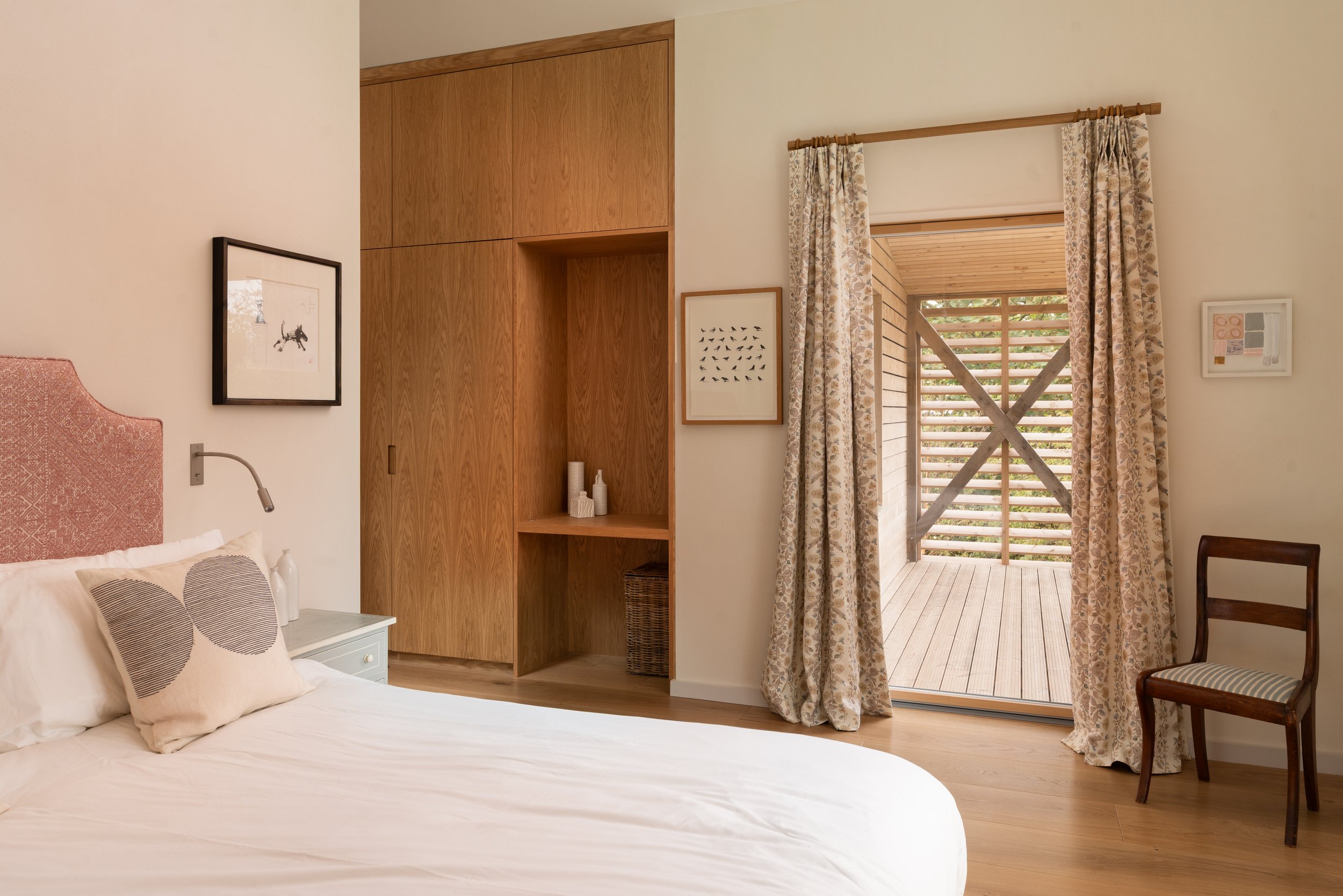 A bedroom with a white bed, pillows, and artwork on the walls. There is wooden cabinetry and a view outside through a sliding door with floral curtains.