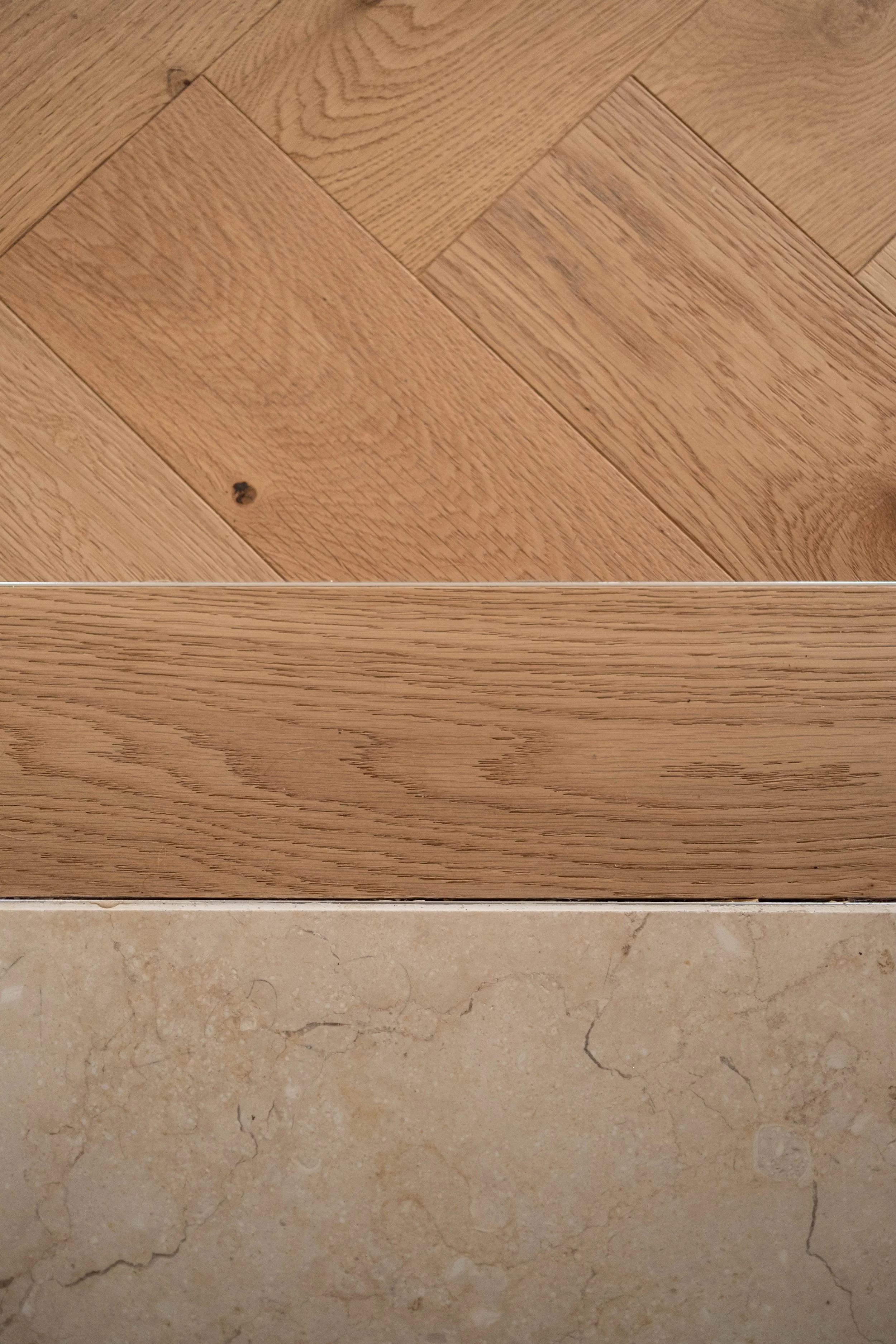 Close-up of a wooden floor with a diagonal pattern, a section of a wooden cabinet or furniture with a horizontal grain, and beige marble or stone flooring with natural veining.