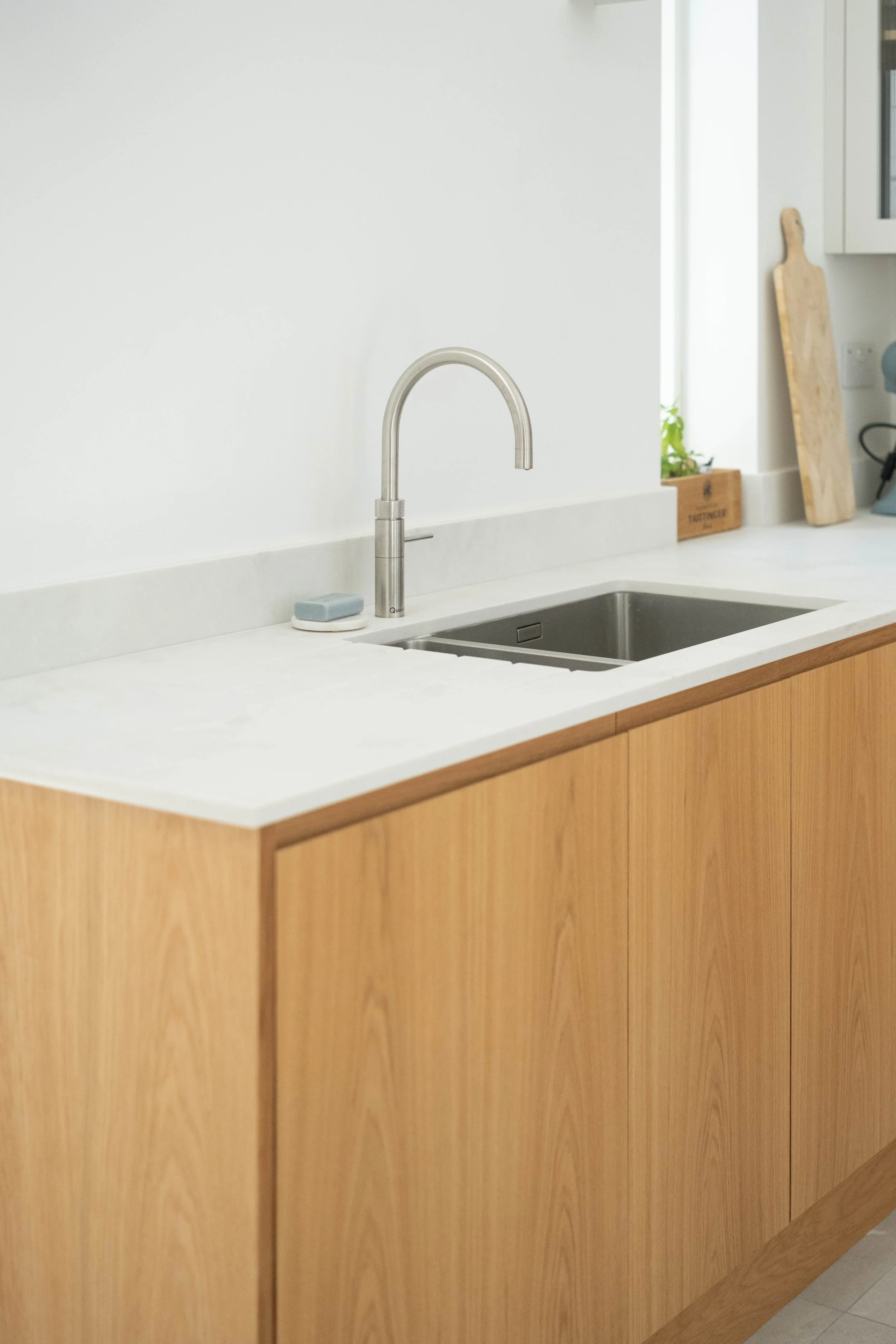 Modern kitchen with white countertop, stainless steel sink, soap, wooden cutting boards, small plant, and cabinetry.