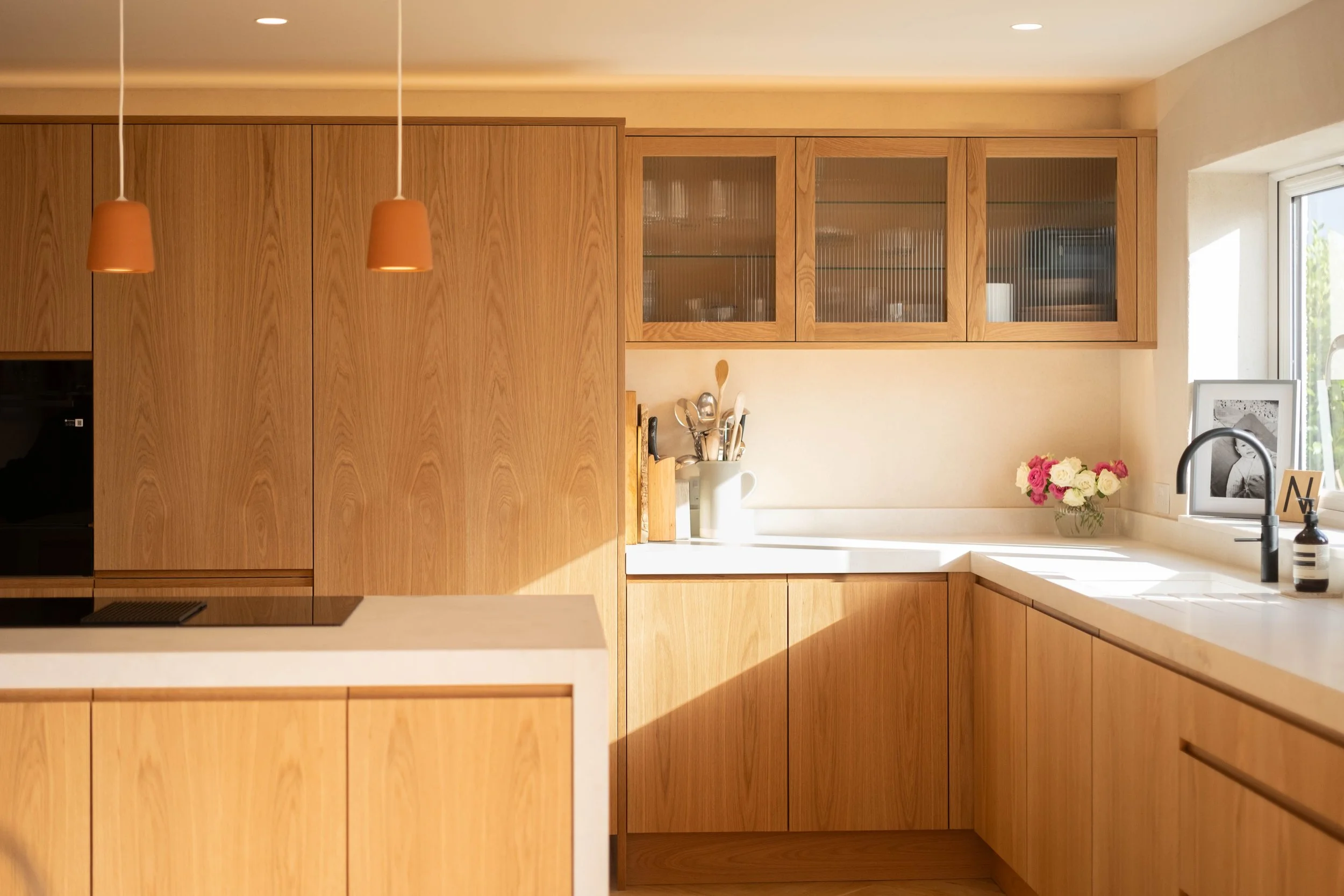 Modern kitchen with wooden cabinets, white countertop, and a window with sunlight. Decor includes a vase with pink and white flowers, framed photo, and kitchen utensils.
