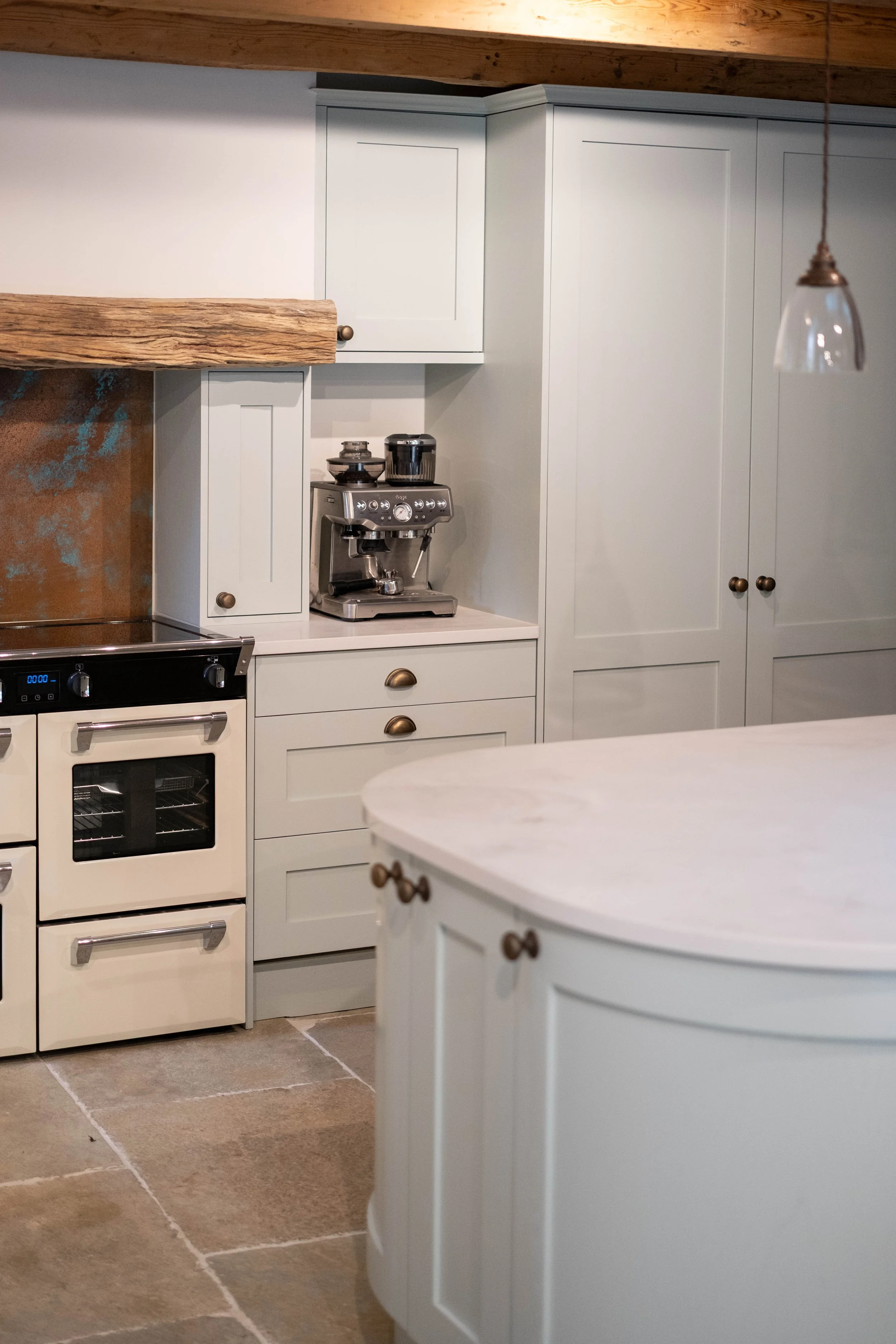A kitchen with white cabinets, a beige oven, and a coffee machine on the counter. The floor has large beige tiles, and a wooden ceiling beam is visible.