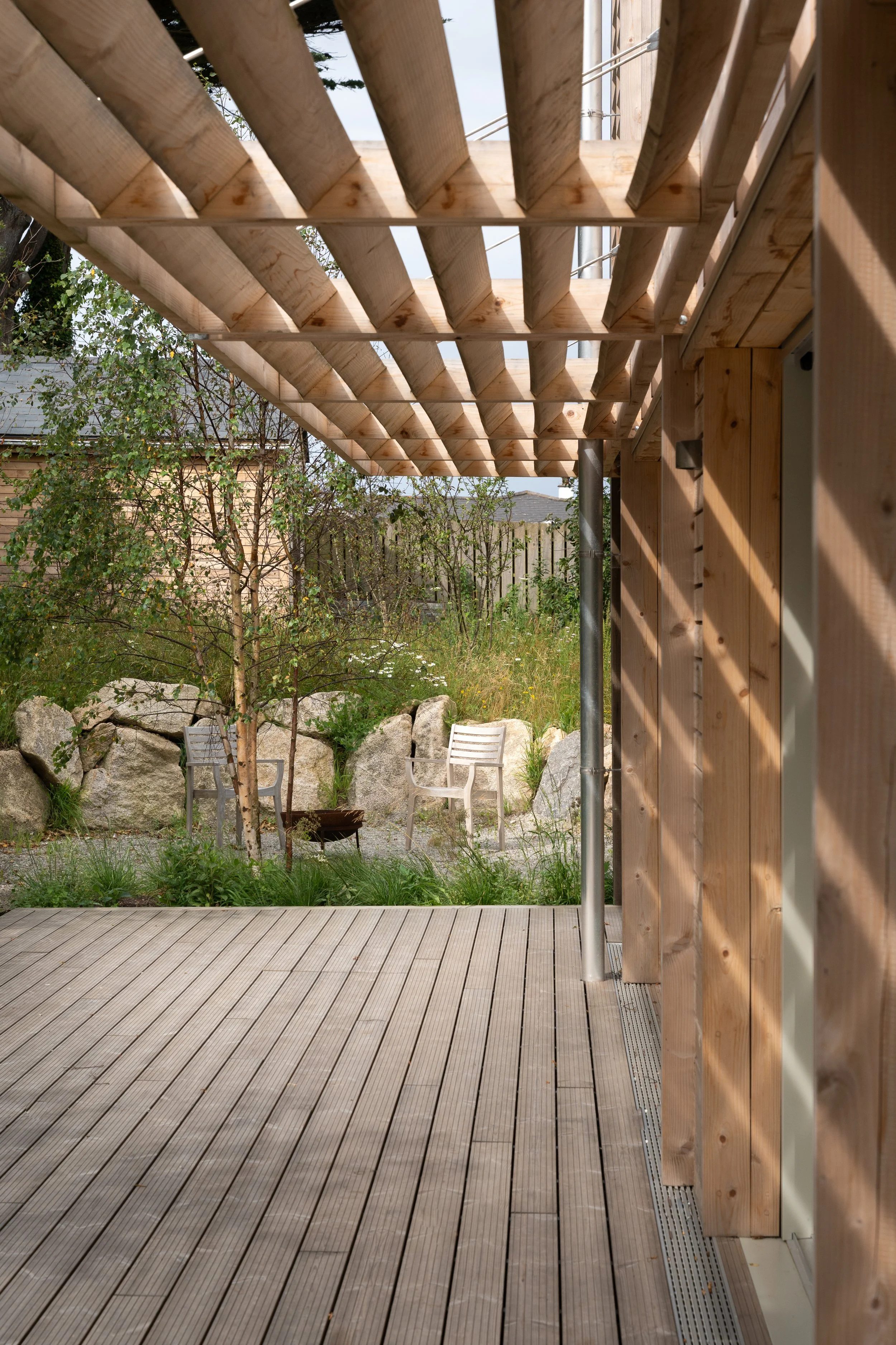 Wooden pergola with slats, a small garden area with two chairs and a young tree, rocks, and greenery in the background.