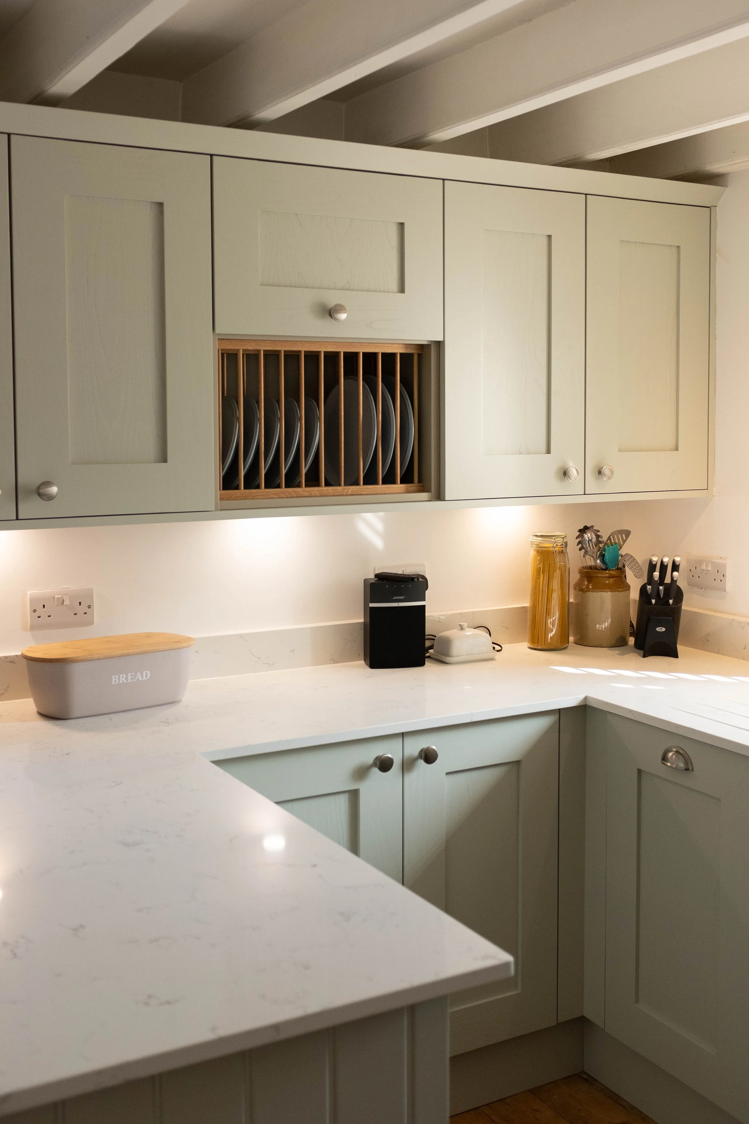 A kitchen with pale green cabinets, white countertops, a dish rack with plates, and various jars and utensils on the counter.