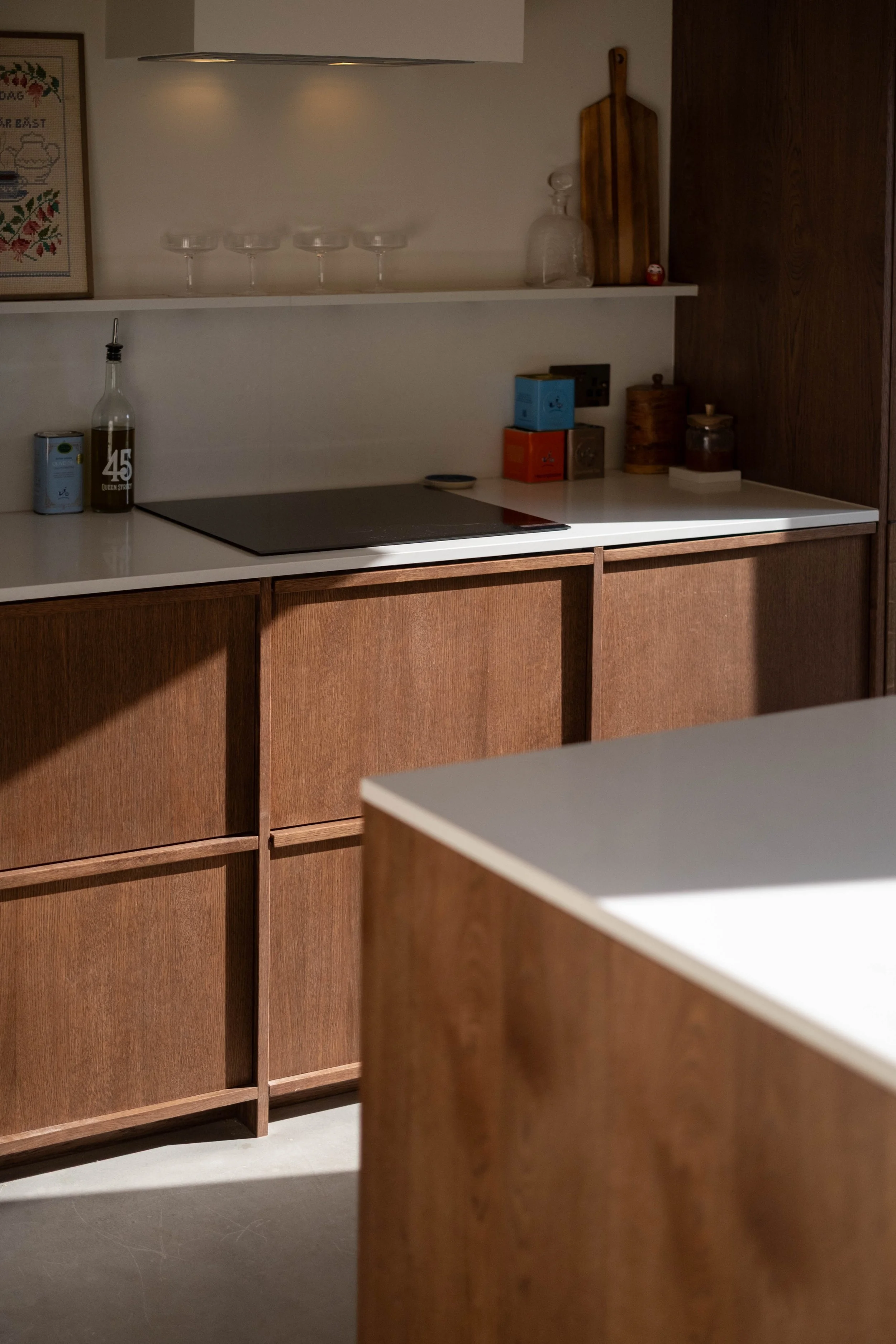 A modern kitchen with wooden cabinets, white countertops, and a built-in electric cooktop. The upper open shelf displays four empty cocktail glasses, a framed cross-stitch artwork, and some glass bottles, including a decanter. The lower cabinet area 