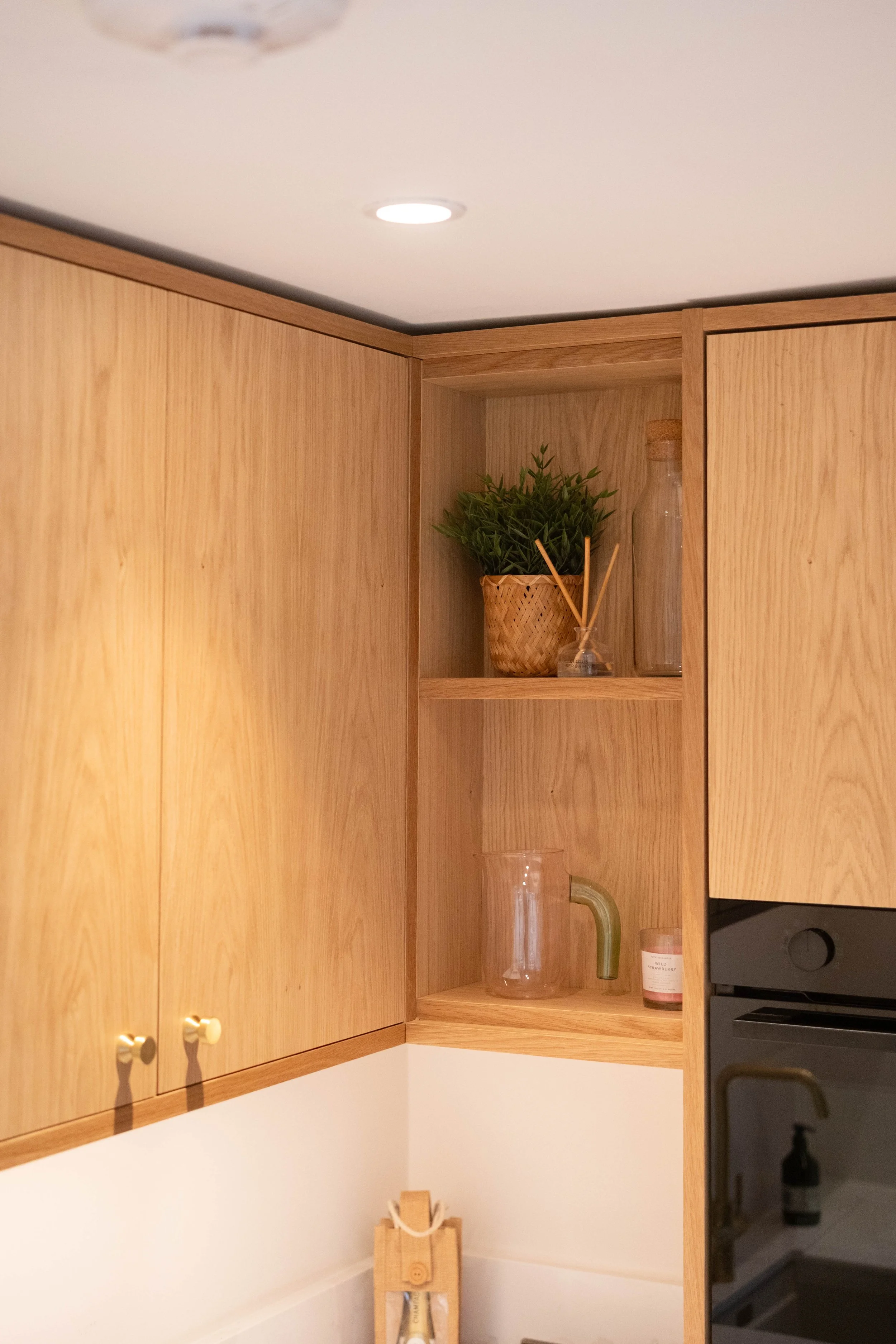 Wooden kitchen cabinets with open shelves holding a potted plant, glass bottles, and small jars; a white counter below and a black appliance to the right.