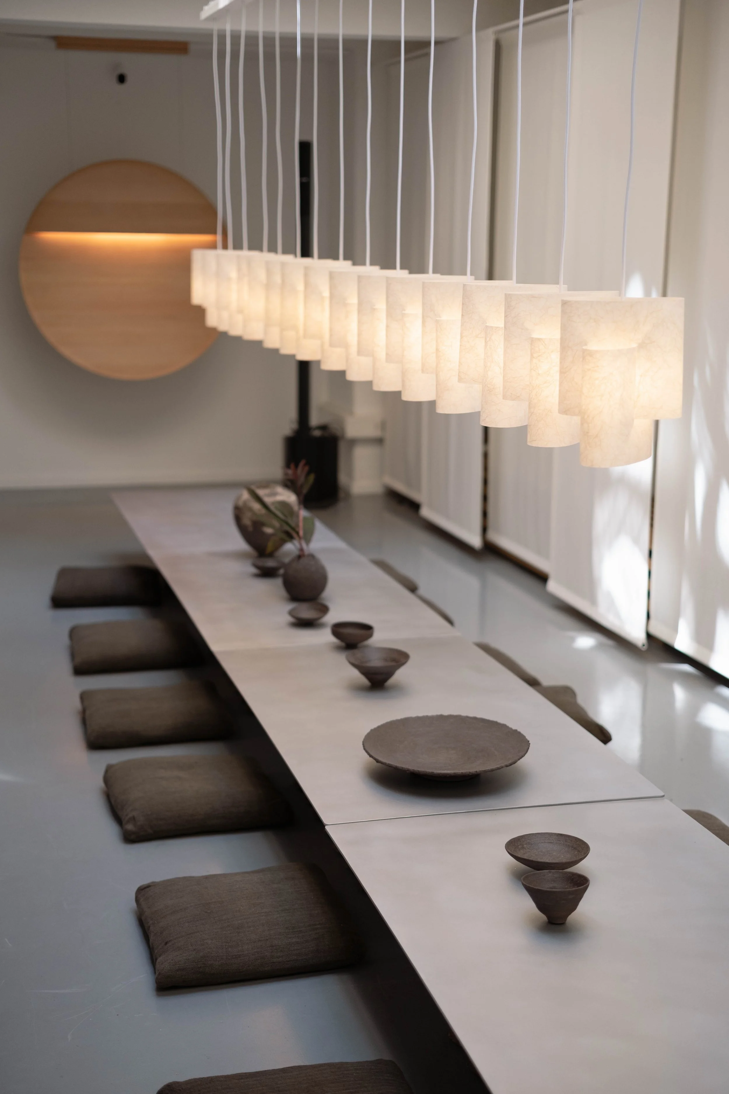A modern dining room with a long white table, brown cushions, and beige bowls. Ceiling hanging pendant lights provide warm illumination, and a decorative wooden wall feature is visible in the background.