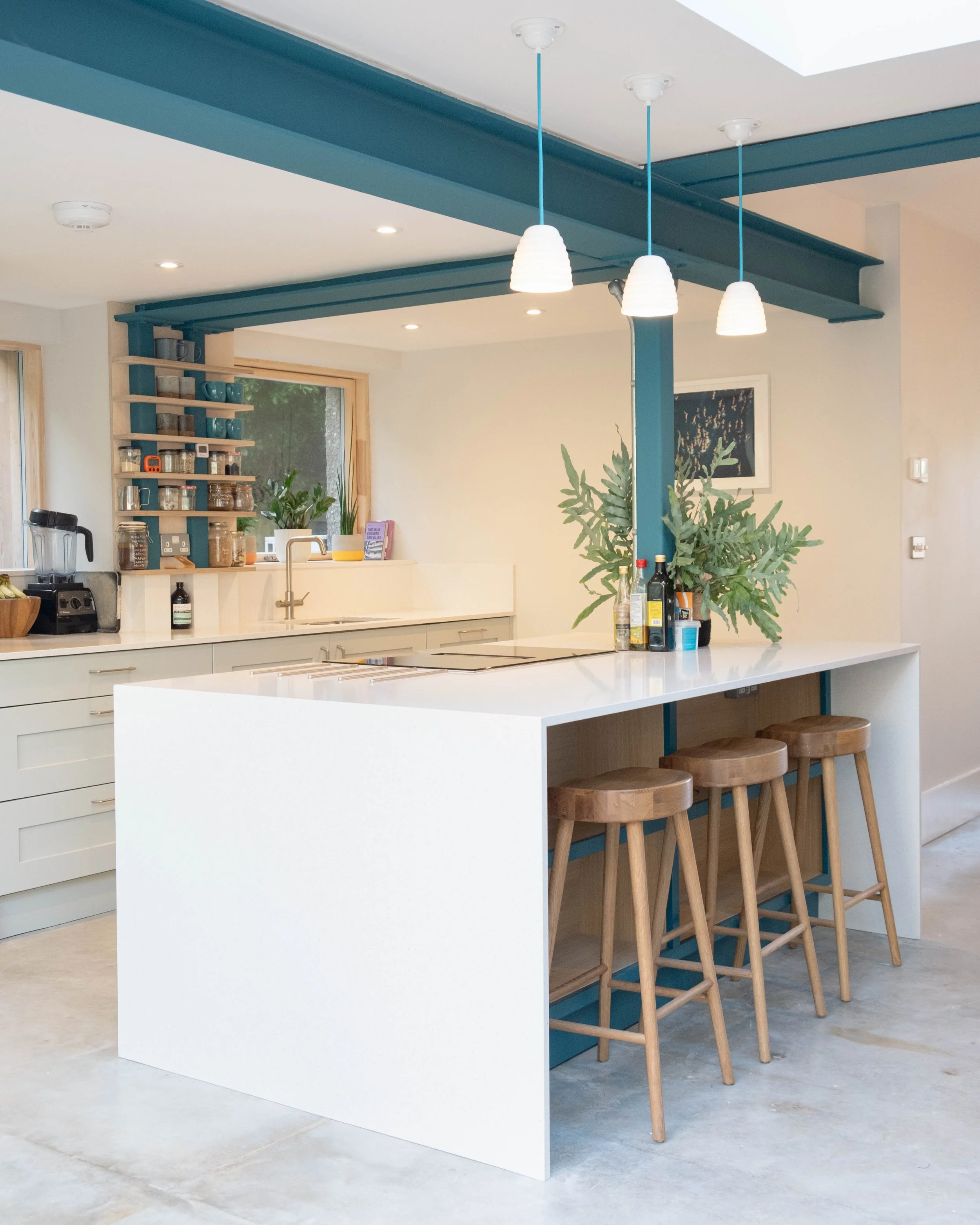 Modern kitchen with white island, four wooden barstools, blue support beams, open shelves with jars and cups, potted plants, and pendant lights.