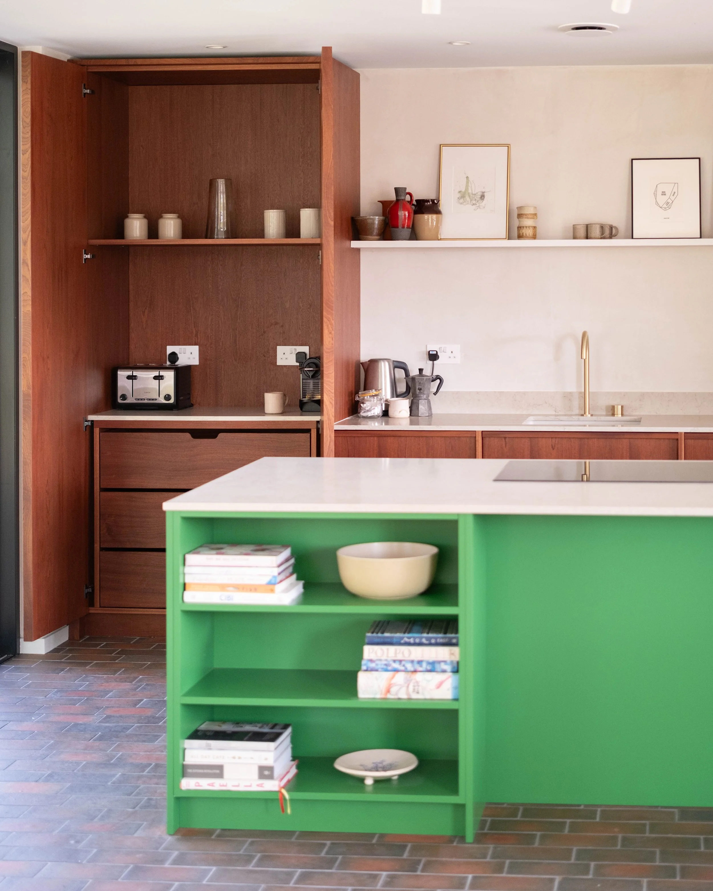 A kitchen area with a green bookshelf displaying books and dishes in the foreground, and a wooden cabinet with decorative vases and a toaster in the background, adjacent to a white wall with artwork and kitchen appliances on the countertop.