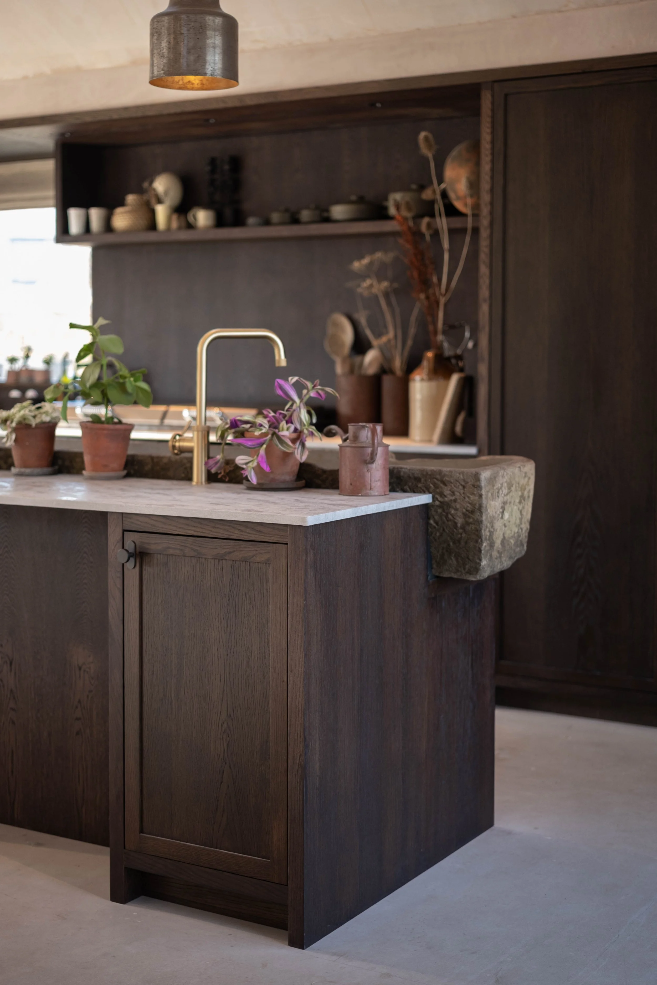 A wooden kitchen island with a marble countertop and a brass faucet. Behind it, dark wooden cabinets and open shelves with dishes, vases, and dried plants. Natural light comes through a window.
