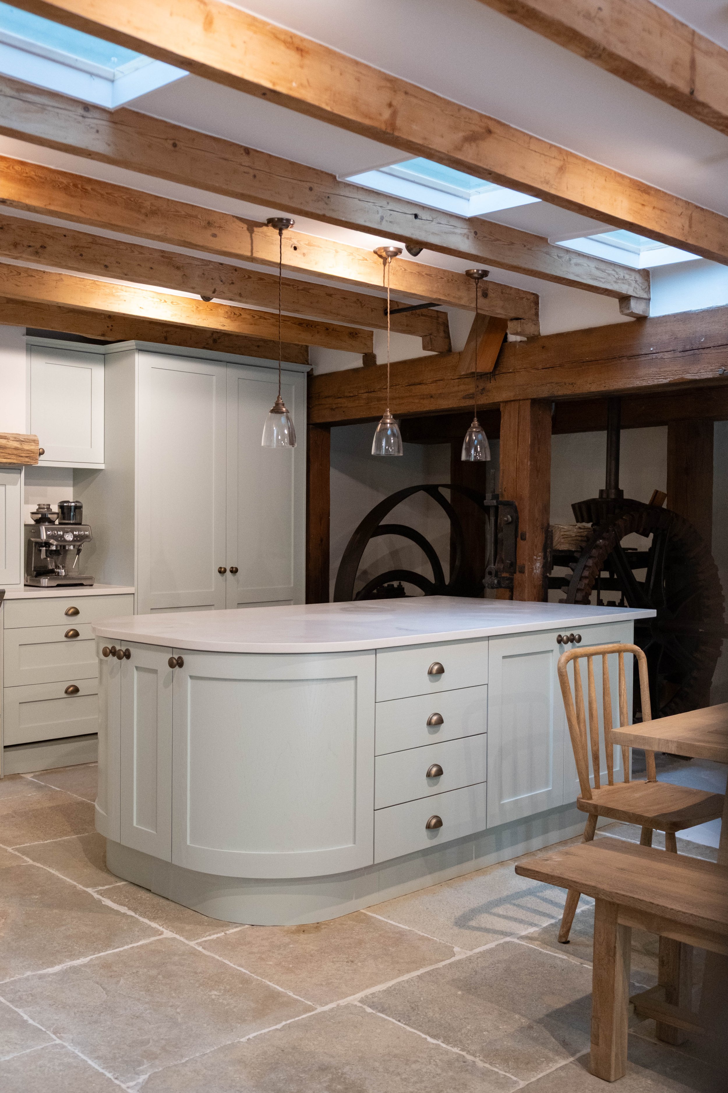 A kitchen with white cabinetry, a kitchen island with a curved front and multiple drawers, three hanging pendant lights, rustic wood beams on the ceiling with skylights, and an exposed wooden beam supporting the ceiling.