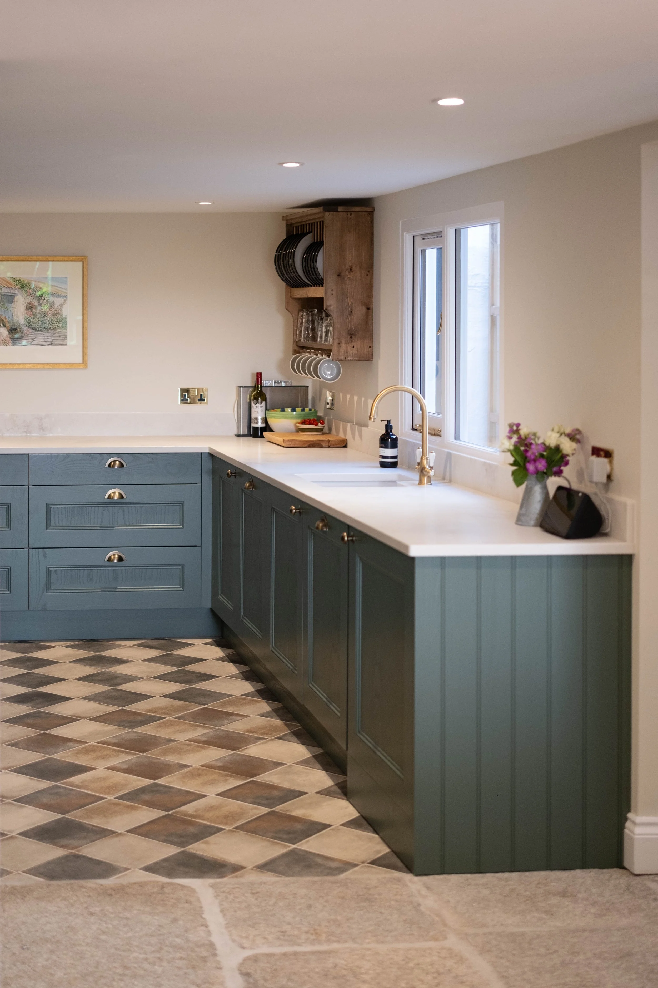 Kitchen with blue cabinets, white countertop, window, and decorative items including a small flower vase, soap dispenser, bottle of wine, bowl of cherries, and a dish towel. The floor has a diamond-patterned tile and a section of beige carpet.