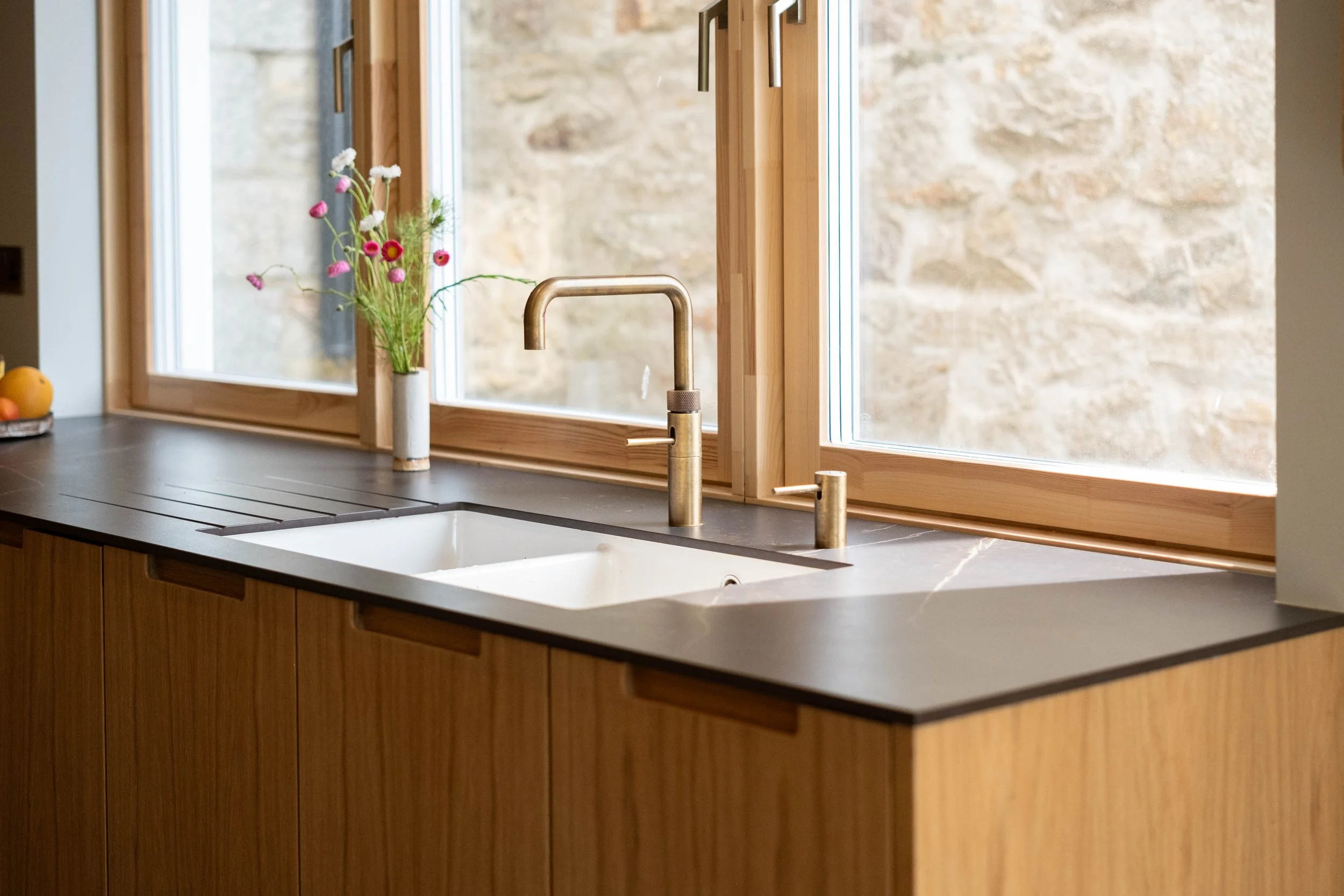 Kitchen sink with a brass faucet in front of a large window with a stone wall outside. A white vase with flowers on the windowsill, and a fruit bowl on the countertop.