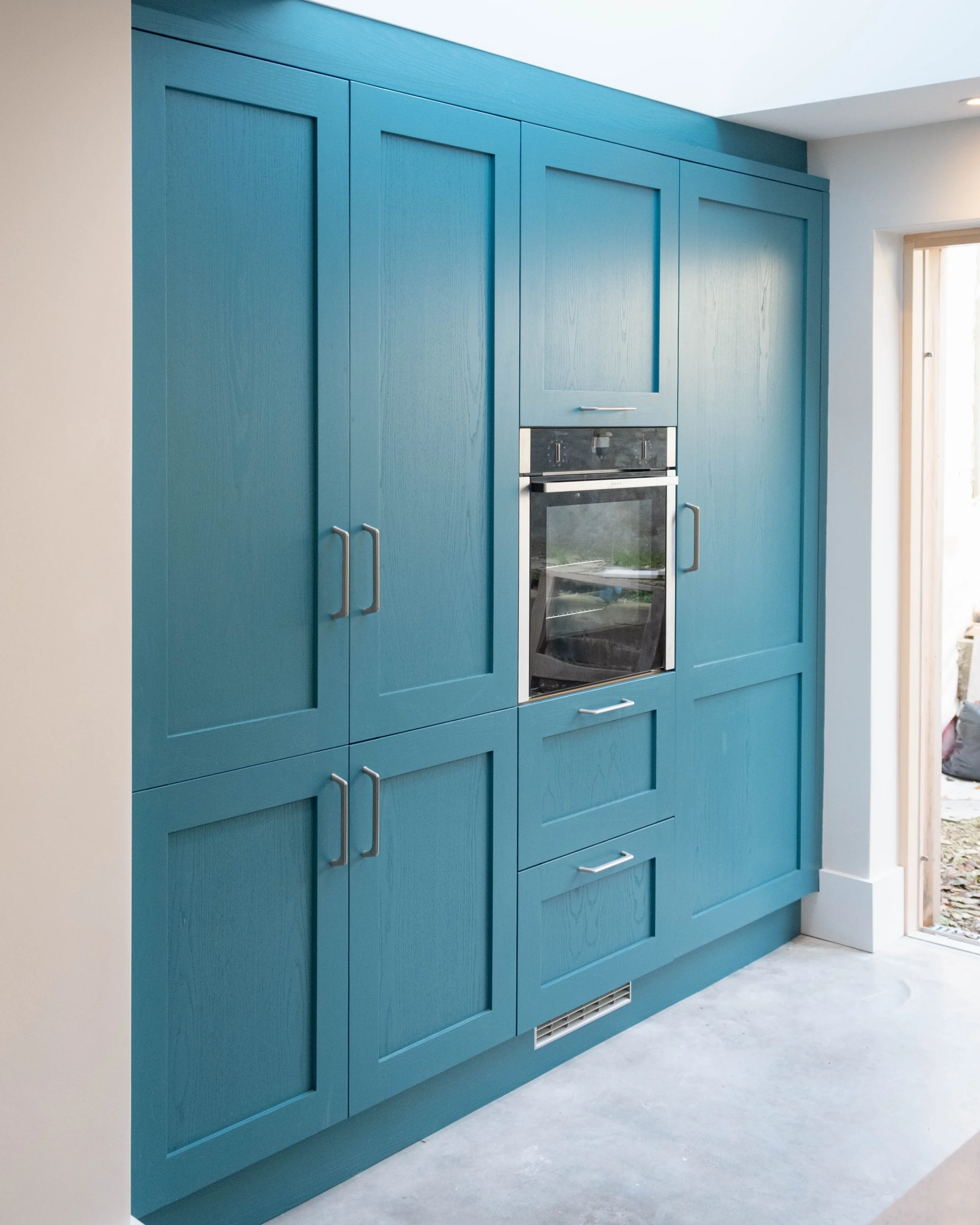 Interior of a kitchen with blue cabinets and built-in oven, next to a door or window with natural light.