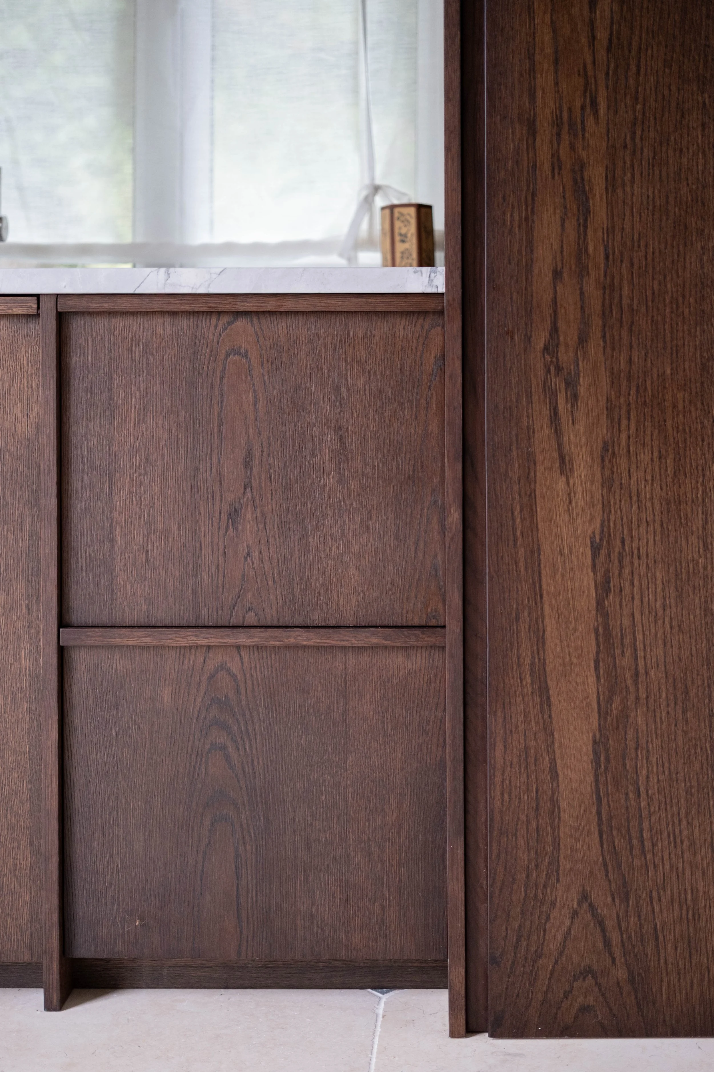 Close-up of a wooden cabinet with a marble countertop, against a background of a window with sheer curtains, and a small book on the windowsill.