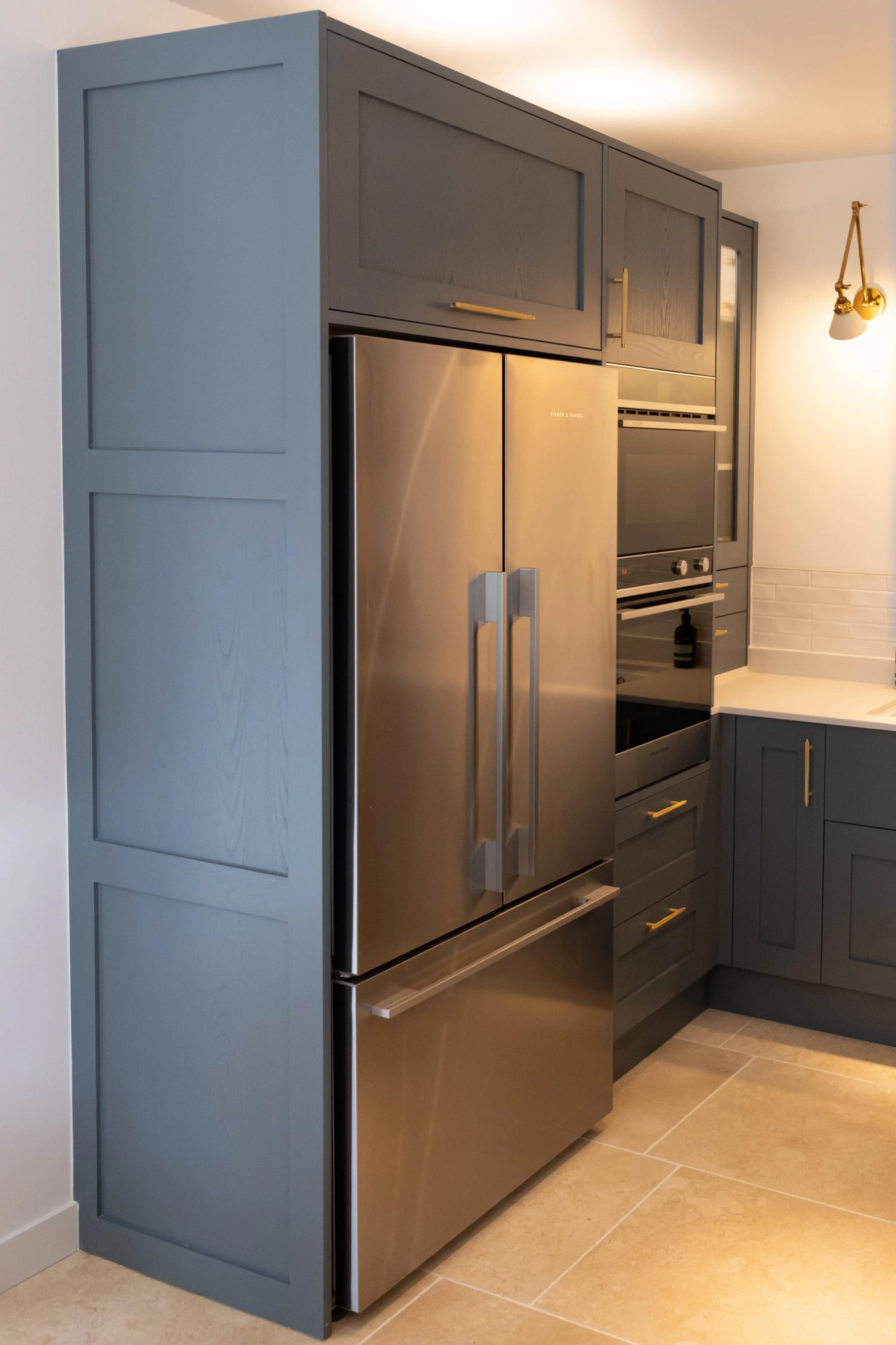 A kitchen with blue cabinets, a stainless steel refrigerator, and built-in oven and microwave. The floor is tiled in beige.