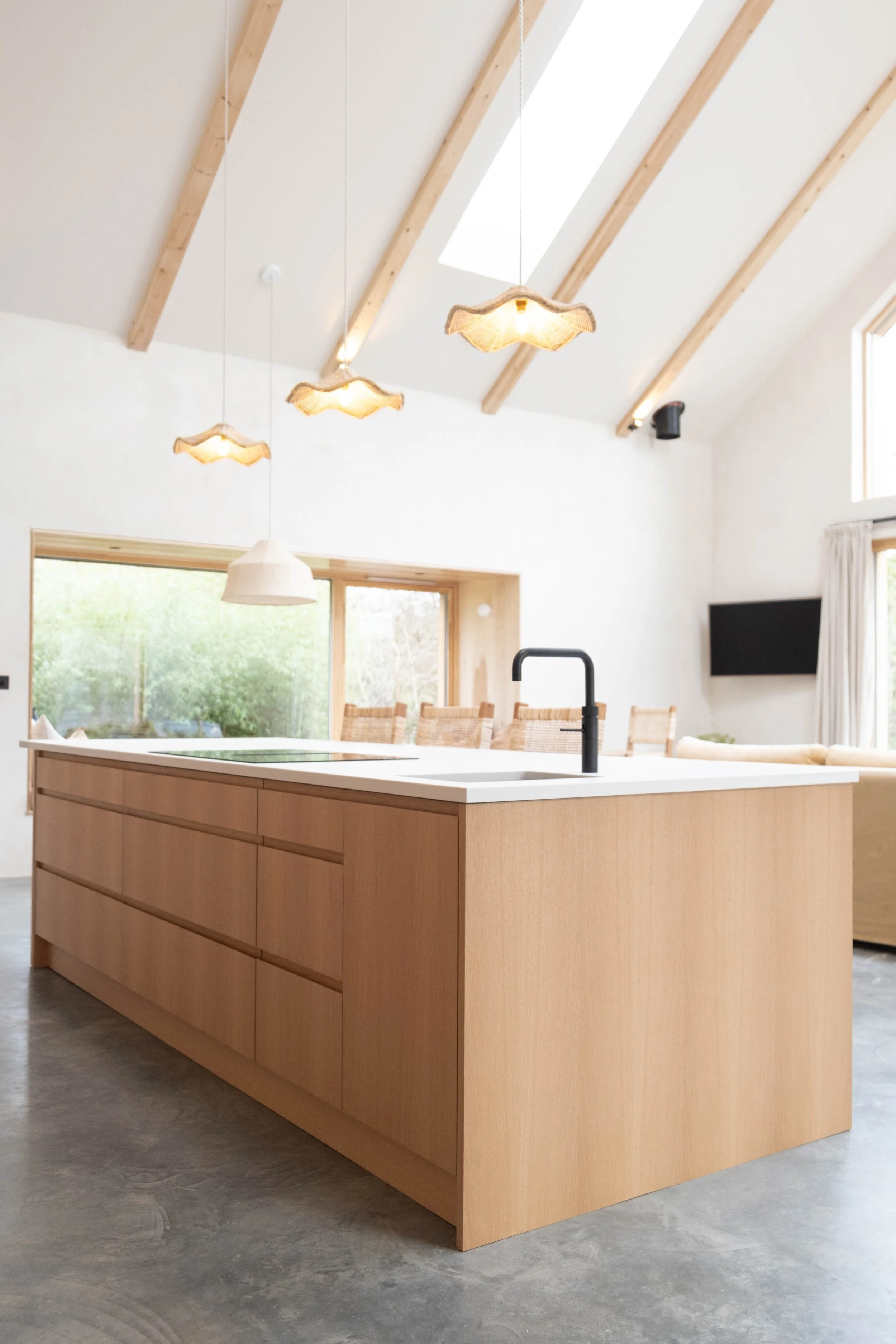A modern kitchen with a large wooden island, black faucet, and pendant lights with wavy shaped shades hanging from a wooden ceiling with skylights. In the background, there are large windows with views of greenery and a mounted flat-screen TV.