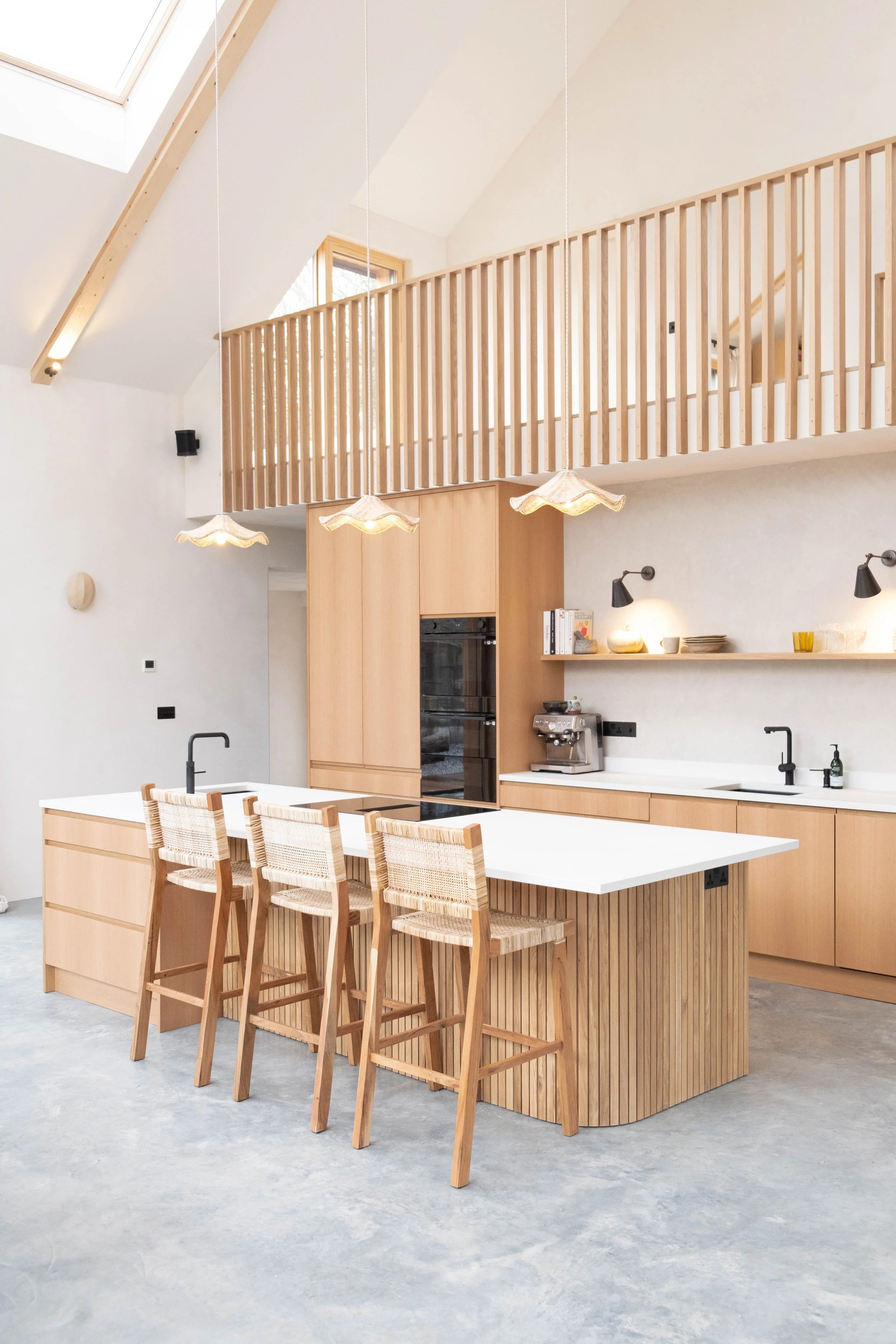Modern kitchen with a large white island, four wooden chairs with woven seats, wooden cabinetry, and a high ceiling with skylights.