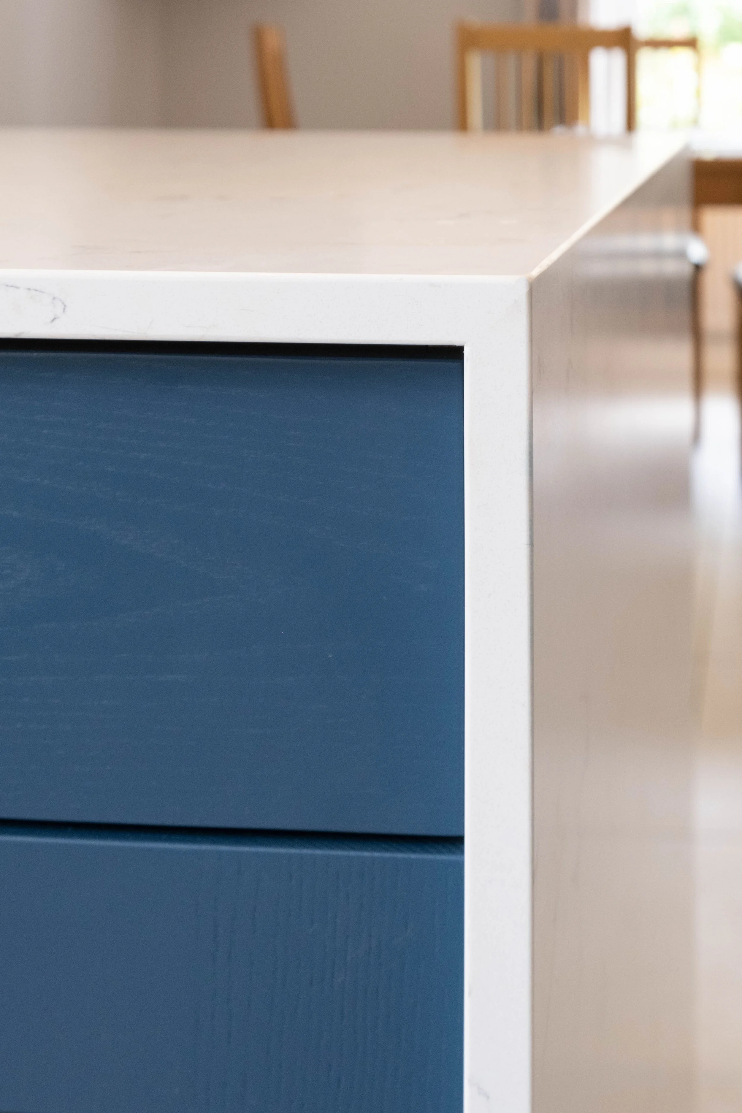 Close-up of a portion of a kitchen island or table with a white marble top and a blue drawer or cabinet front, with a dining area with wooden chairs visible in the background.