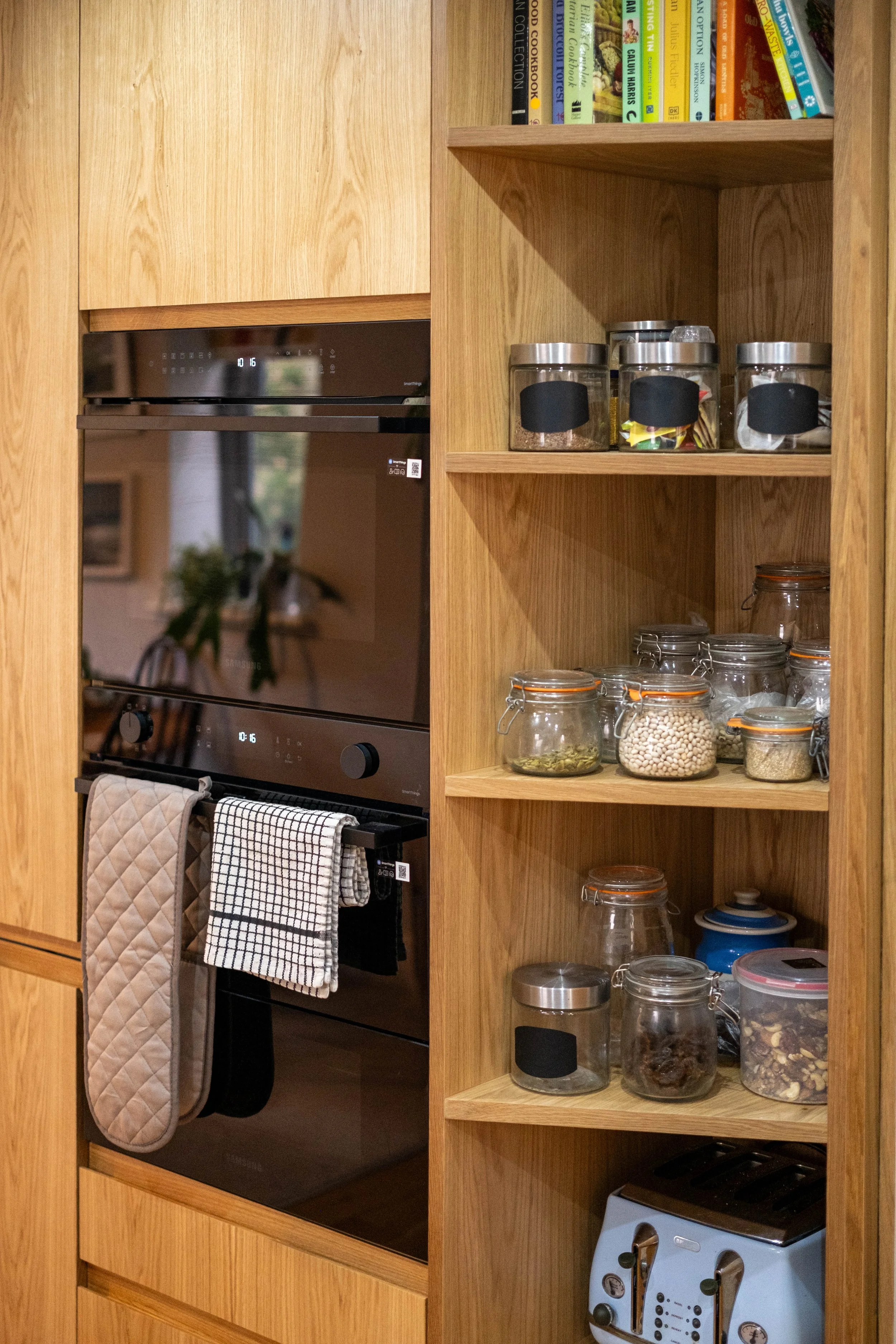 Kitchen with wooden cabinetry, a black oven with a digital clock, kitchen towels hanging on the oven handle, open shelves with glass jars filled with spices, beans, dried herbs, and other ingredients, a toaster oven on the bottom shelf, a potted plan