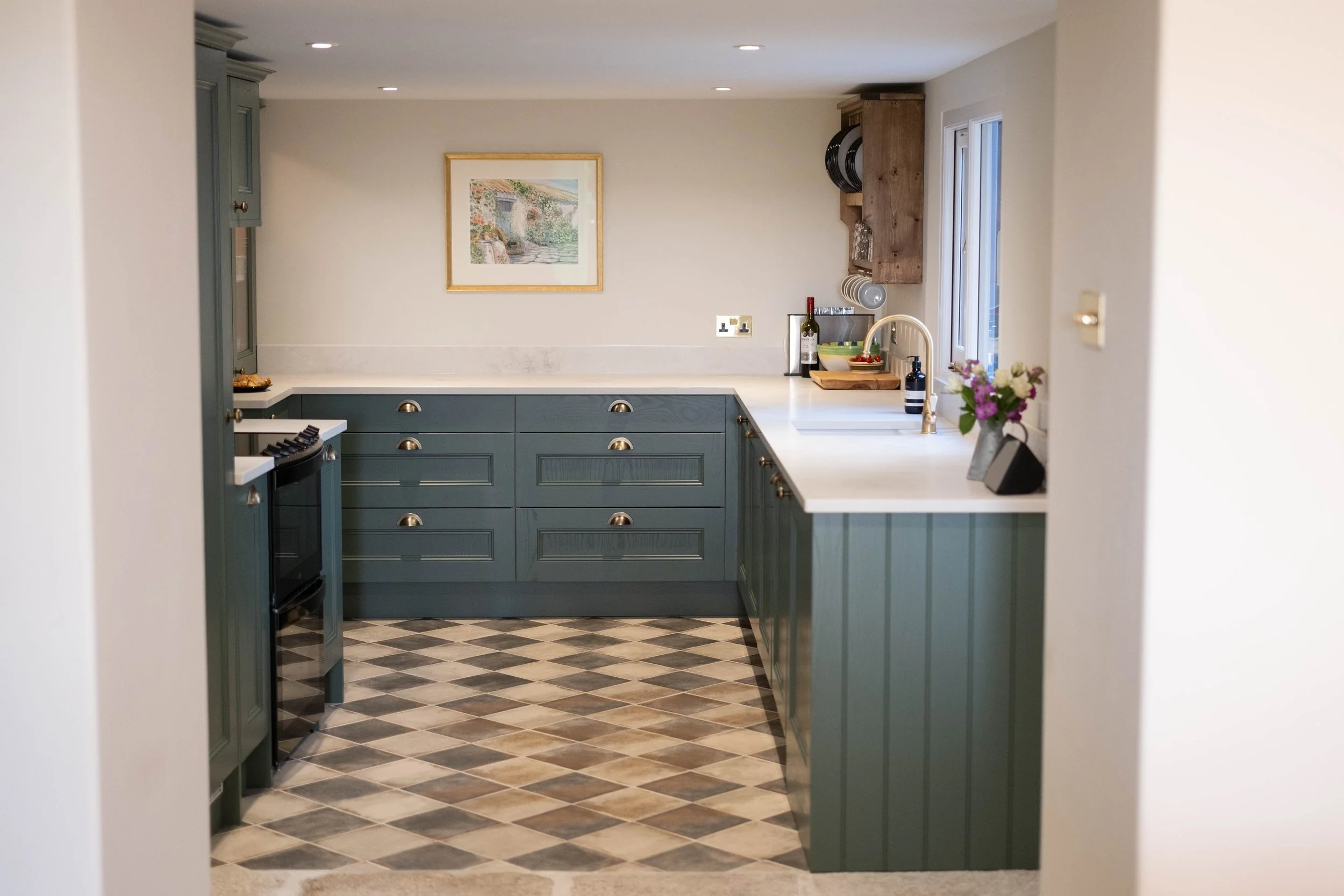 Kitchen with green cabinets, white countertops, patterned tile floor, and a window above the sink, decorated with a flower vase and wine bottle.