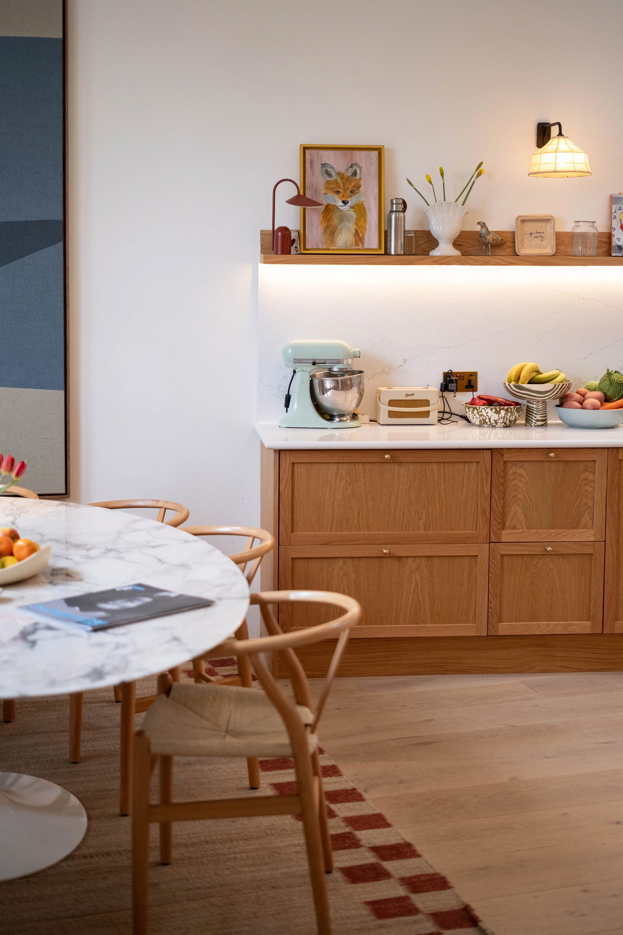 A cozy kitchen with a wooden countertop, a light blue stand mixer, fruit bowls containing bananas, peaches, and greens, decorative items like a framed fox painting, a vase with tulips, and various small decorative objects. With chairs around a marble