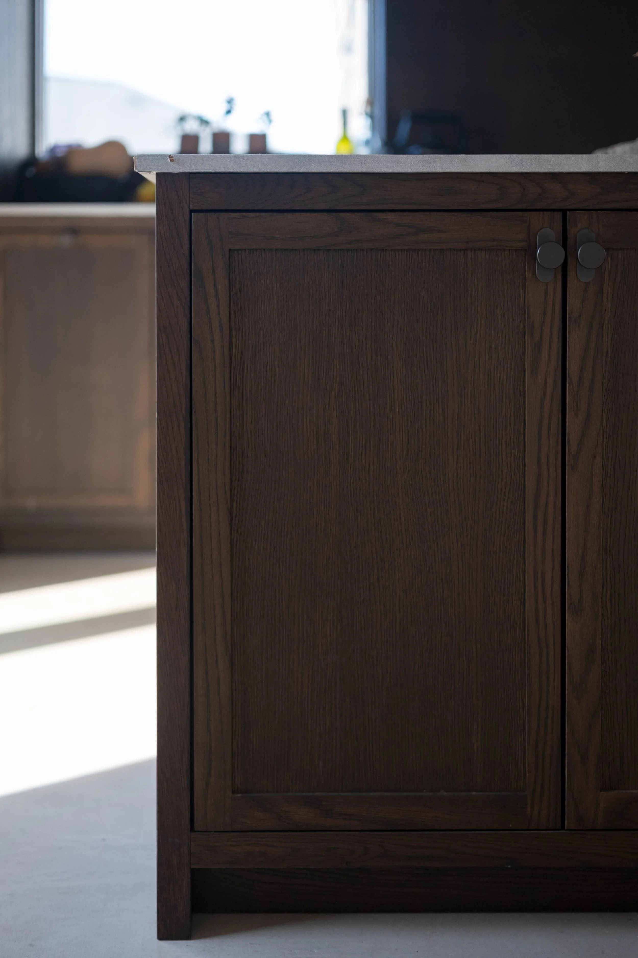 Close-up of a dark wooden cabinet with a light gray countertop, in a well-lit kitchen with a bright window in the background.