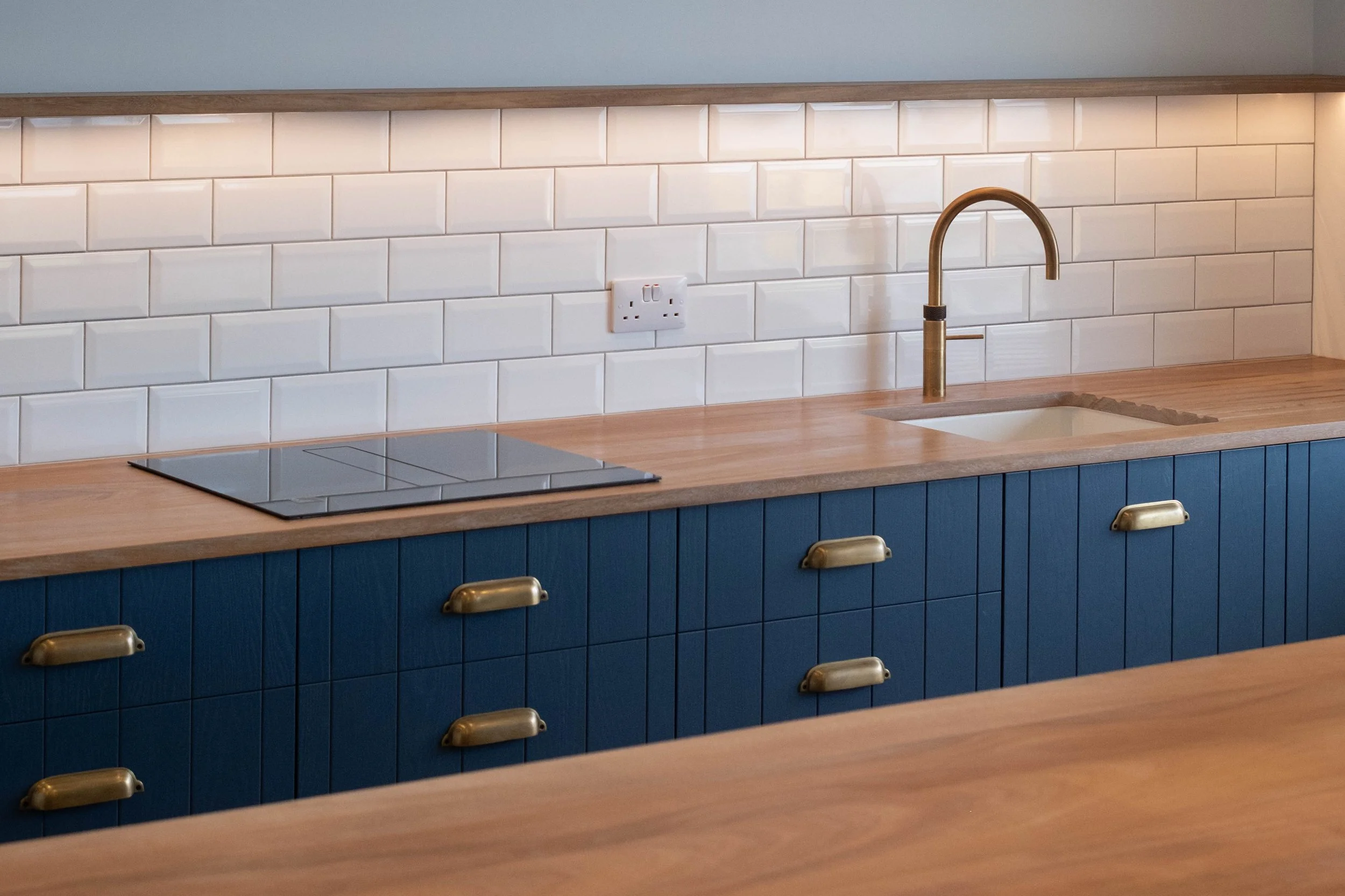 Modern kitchen countertop with a gold sink faucet, white subway tile backsplash, an electric cooktop, a double electrical outlet, and blue cabinets below with brass handles.
