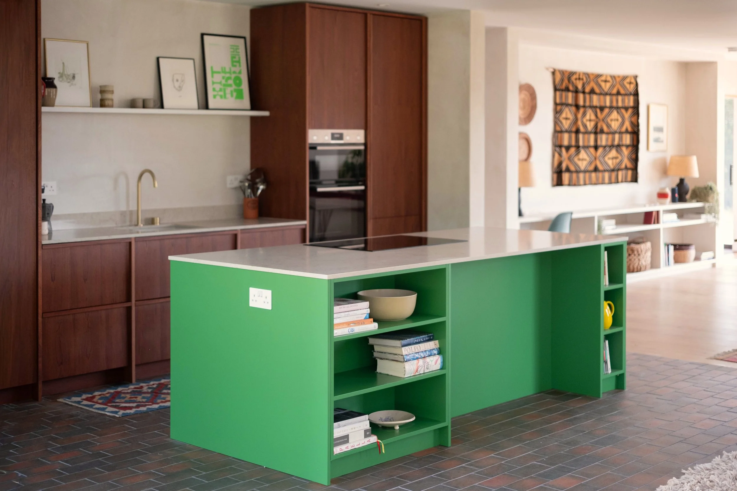 A modern kitchen with green island, wooden cabinets, and a white shelf with framed artwork. Open space with a patterned curtain on the wall and various decorative items.