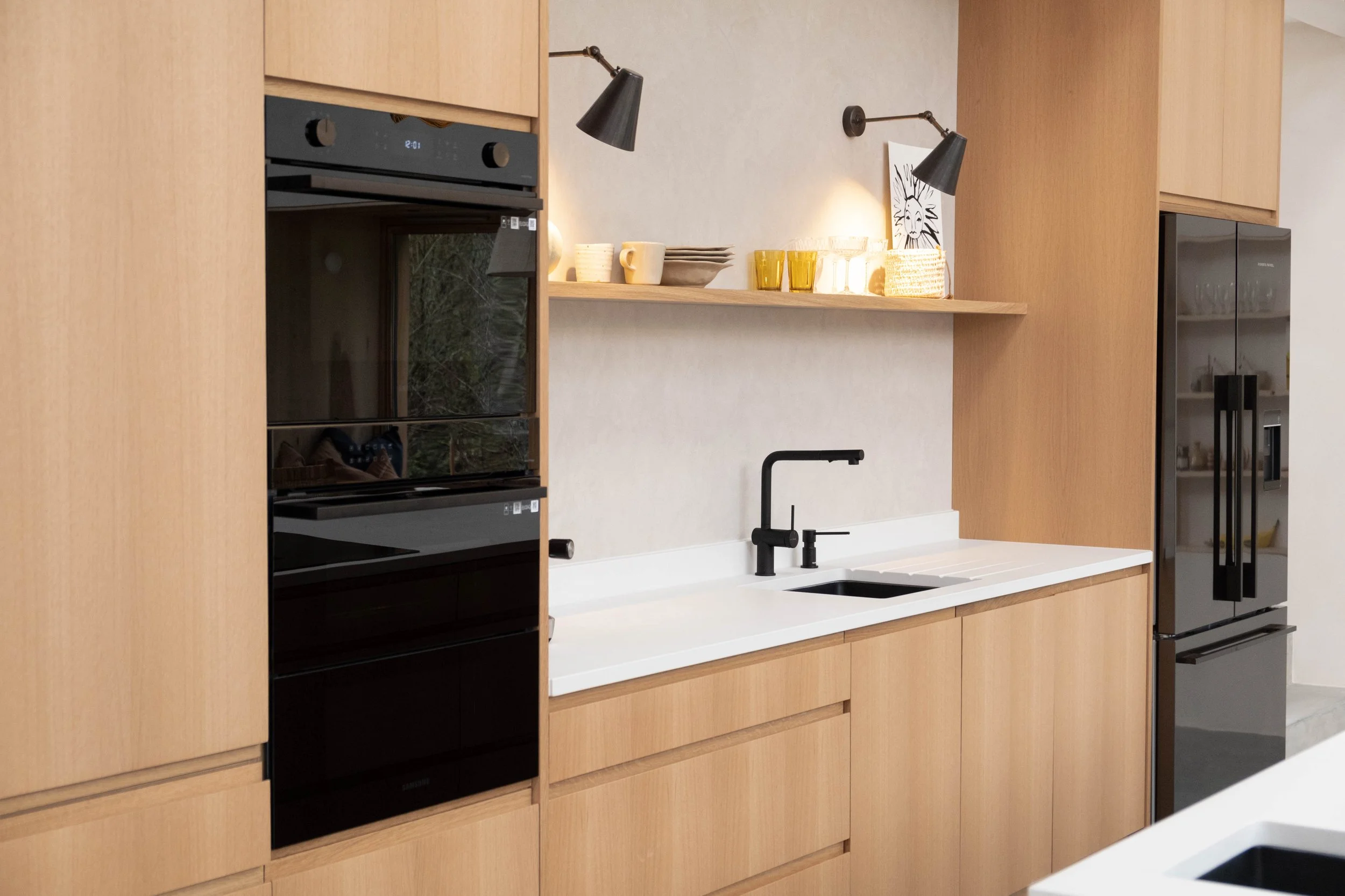 Modern kitchen with wooden cabinetry, black appliances, white countertop, black sink faucet, open shelving with dishes, yellow glasses, and a black and white lion artwork.