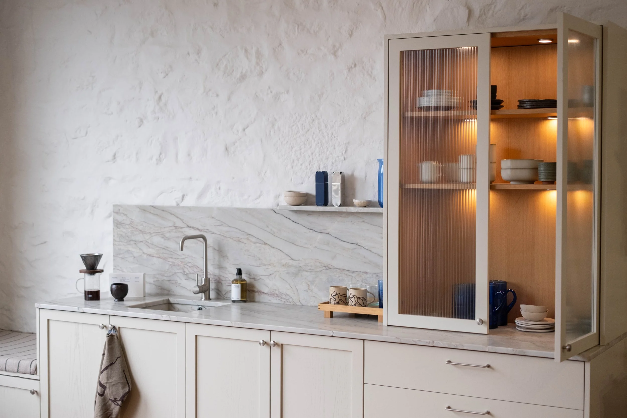 A kitchen with white cabinetry, marble backsplash, and open shelves filled with plates, bowls, and mugs. There is a glass-front cabinet with wooden shelves and warm lighting.