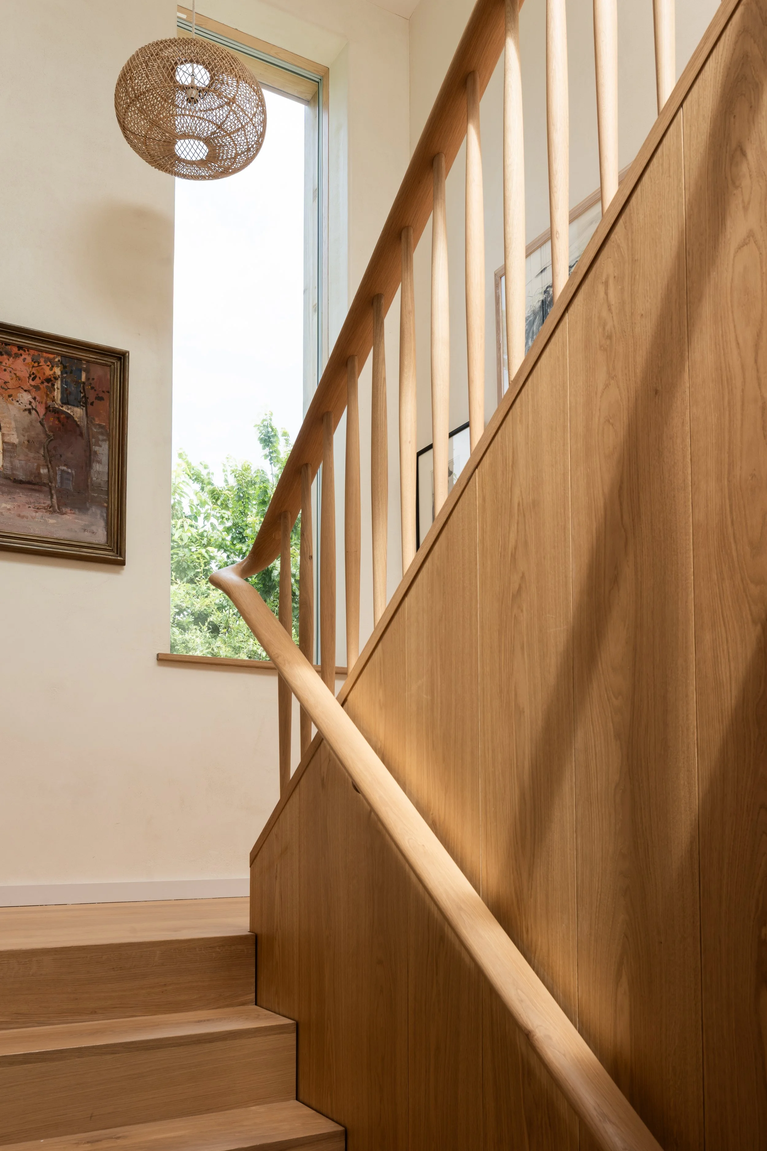 Wood staircase with handrail in a well-lit interior near a large window showing trees and sky outside, with framed artwork on the wall and a woven pendant light with a skull design.
