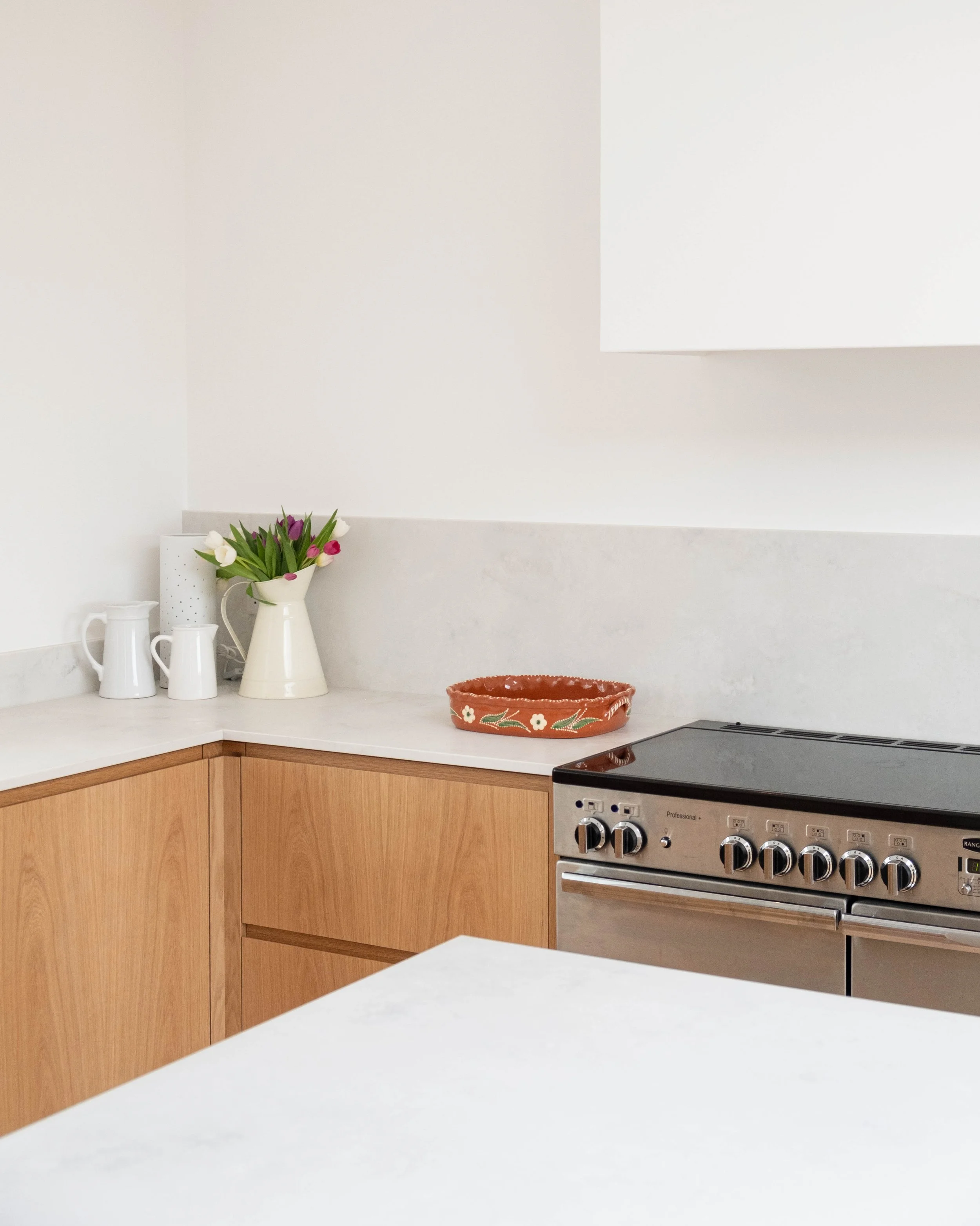 A modern kitchen with a white countertop, wooden cabinet, stainless steel stove, and a white wall. On the counter, there are white pitchers and a vase with tulips. There is a decorative terracotta dish with white flowers painted on it.