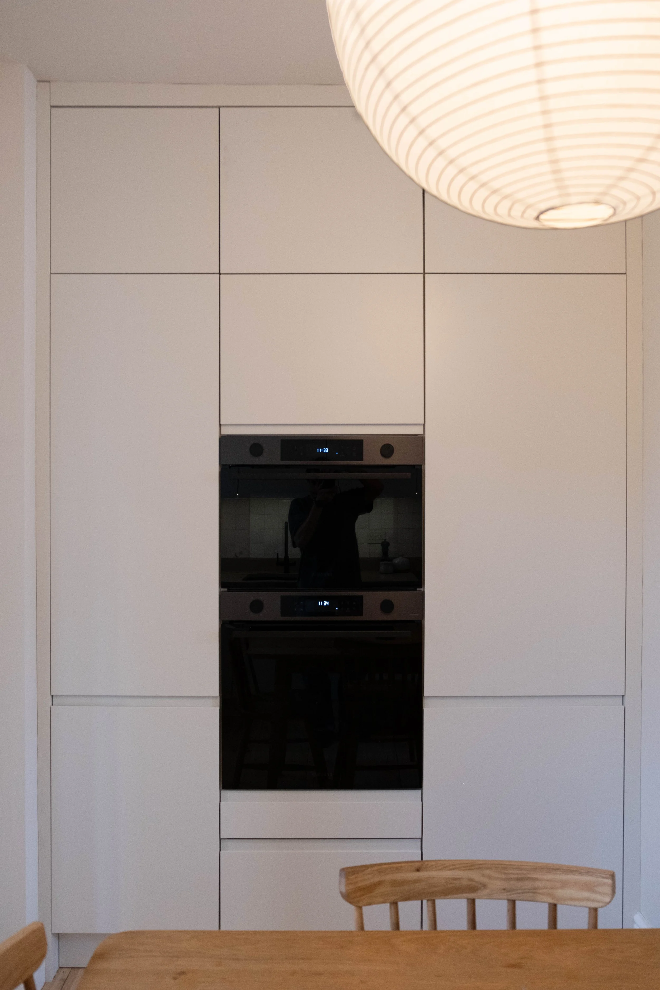 A modern kitchen with white cabinets, a built-in oven and microwave, a wooden dining table with chairs, and a large hanging light fixture.