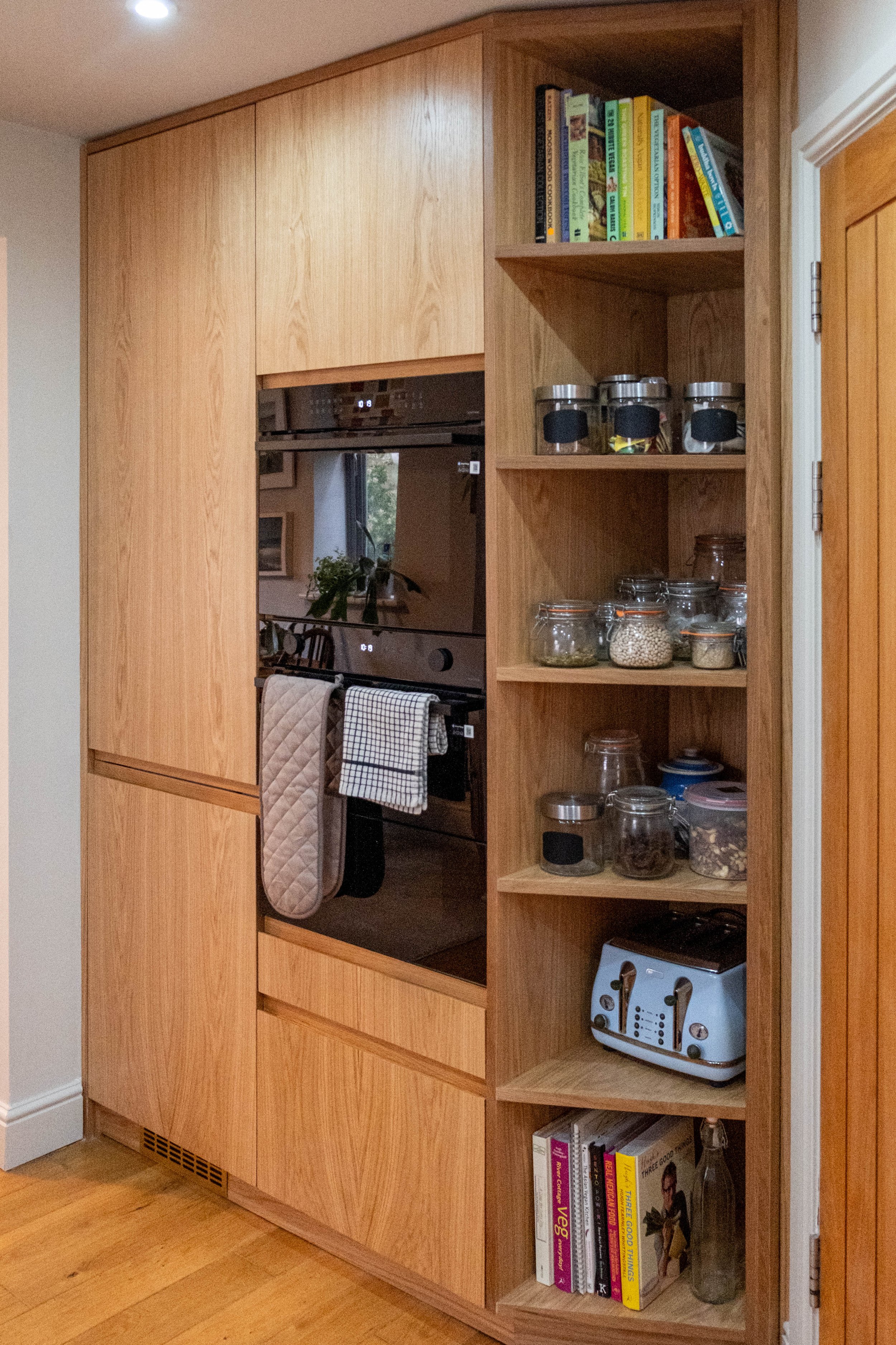 A wooden kitchen cabinet with open shelves displaying cookbooks, jars of food, and a toaster oven. The cabinet has a sleek, modern design with a natural wood finish.